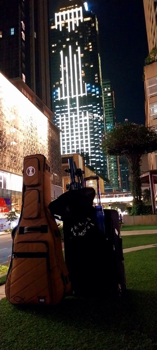 Three golf bags filled with golf clubs are positioned on a grassy area at night. A tall, illuminated modern skyscraper with bright white lights and a decorative facade is seen in the background.
