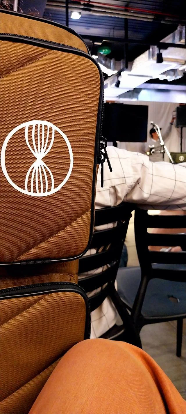 Close-up of a tan backpack with a white logo, sitting on a person's lap at a meeting or event, with a table, chairs, and a person working in the background.