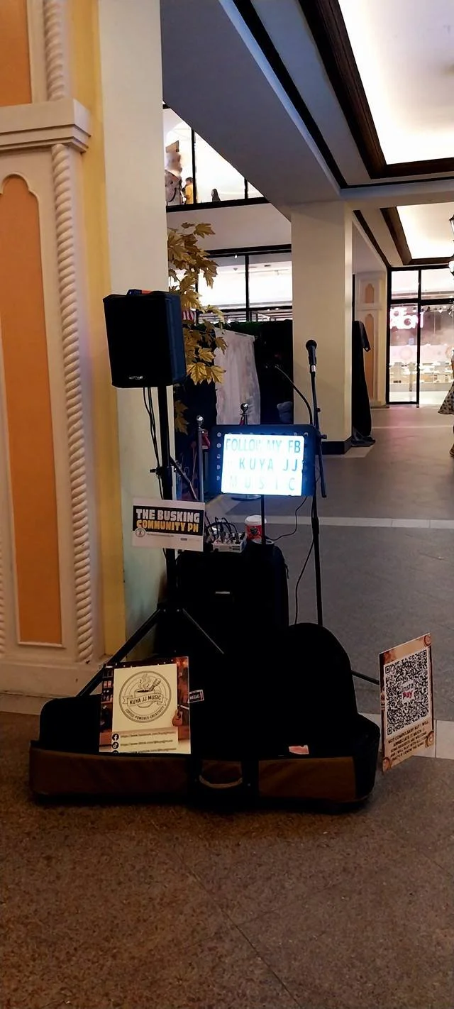 A small music setup with a microphone and speaker in a public indoor space, including a digital sign that reads, 'FOLLOW MY FB KUYA JJ MUSIC,' and promotional signs for the music performer.