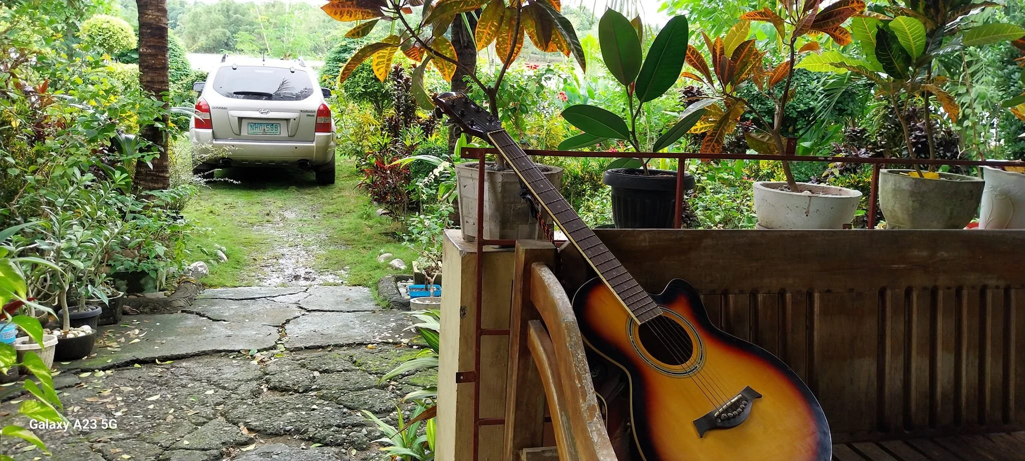 A backyard scene with a silver car parked on a grassy and cracked driveway, surrounded by green plants and trees. An acoustic guitar leans against a wooden railing on a porch decorated with potted plants.