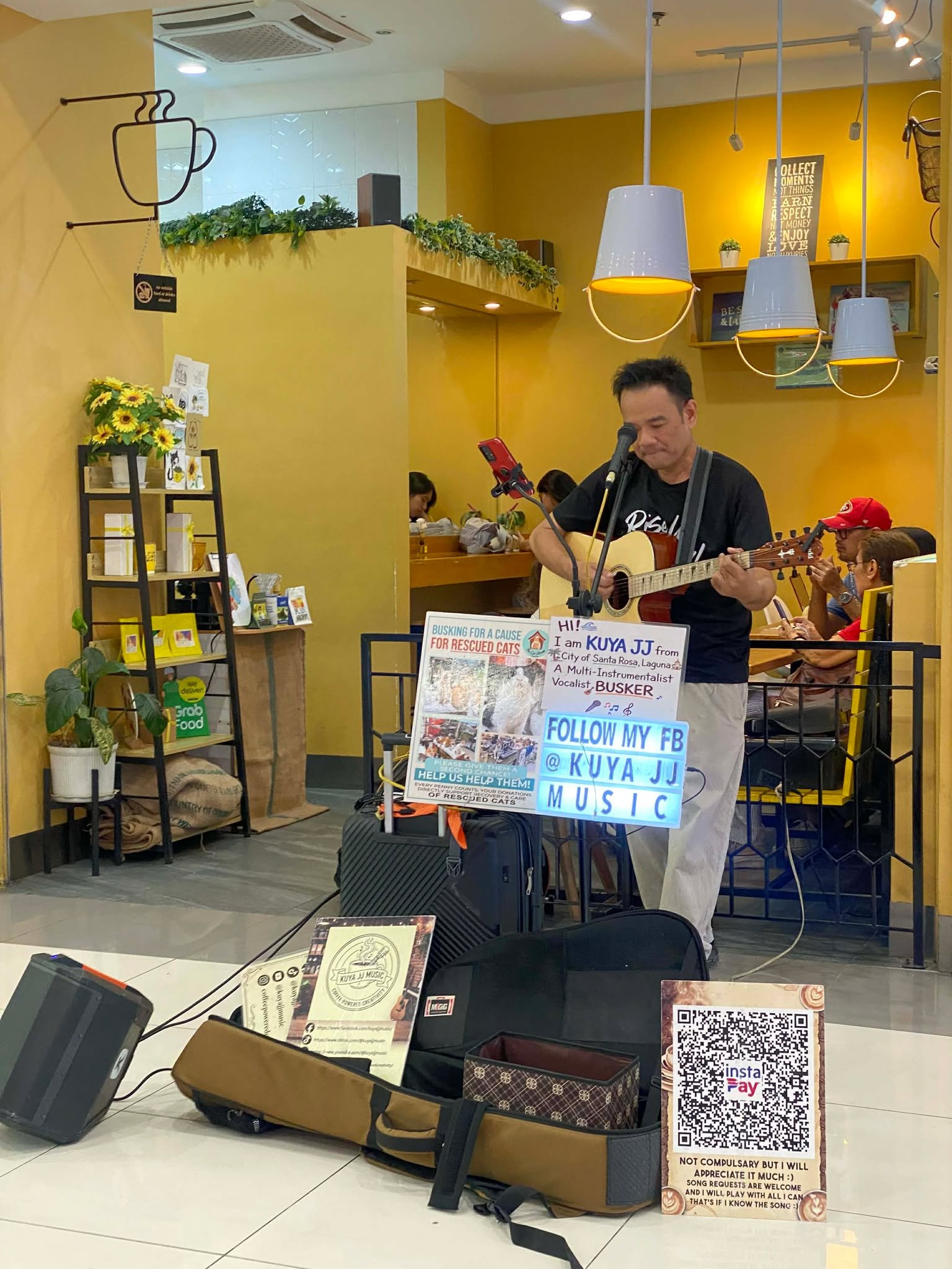 A man is playing guitar and singing into a microphone in a brightly lit cafe. There are teenagers sitting at a table in the background. A large sign in front of the man indicates he is a singer named Kuya JJ from Santa Rosa, Laguna, and mentions supp