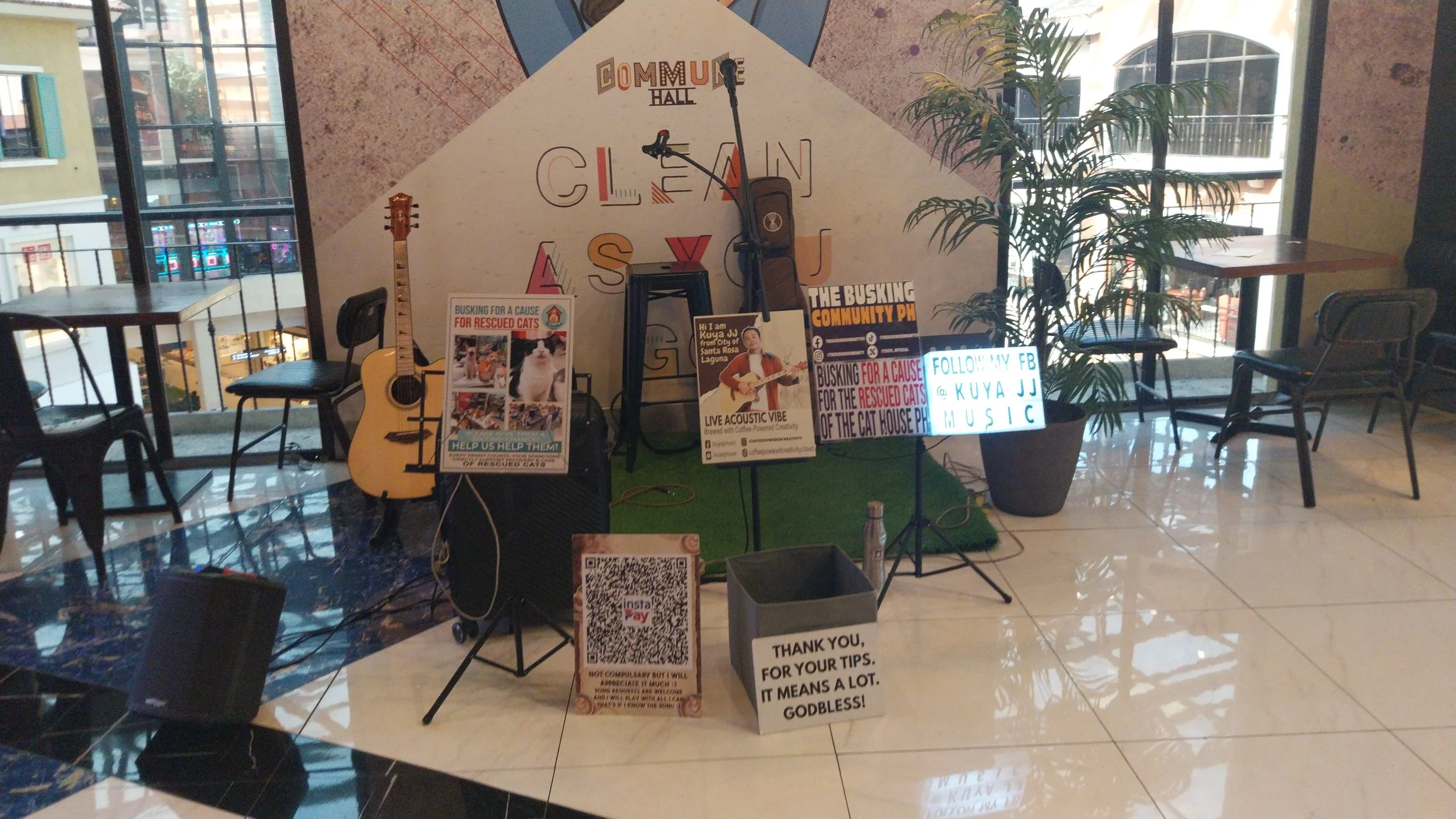 Indoor community event setup with posters, a guitar, microphones, and chairs, near large windows letting in daylight. A wall displays the words 'Community Hall' and other colorful text.