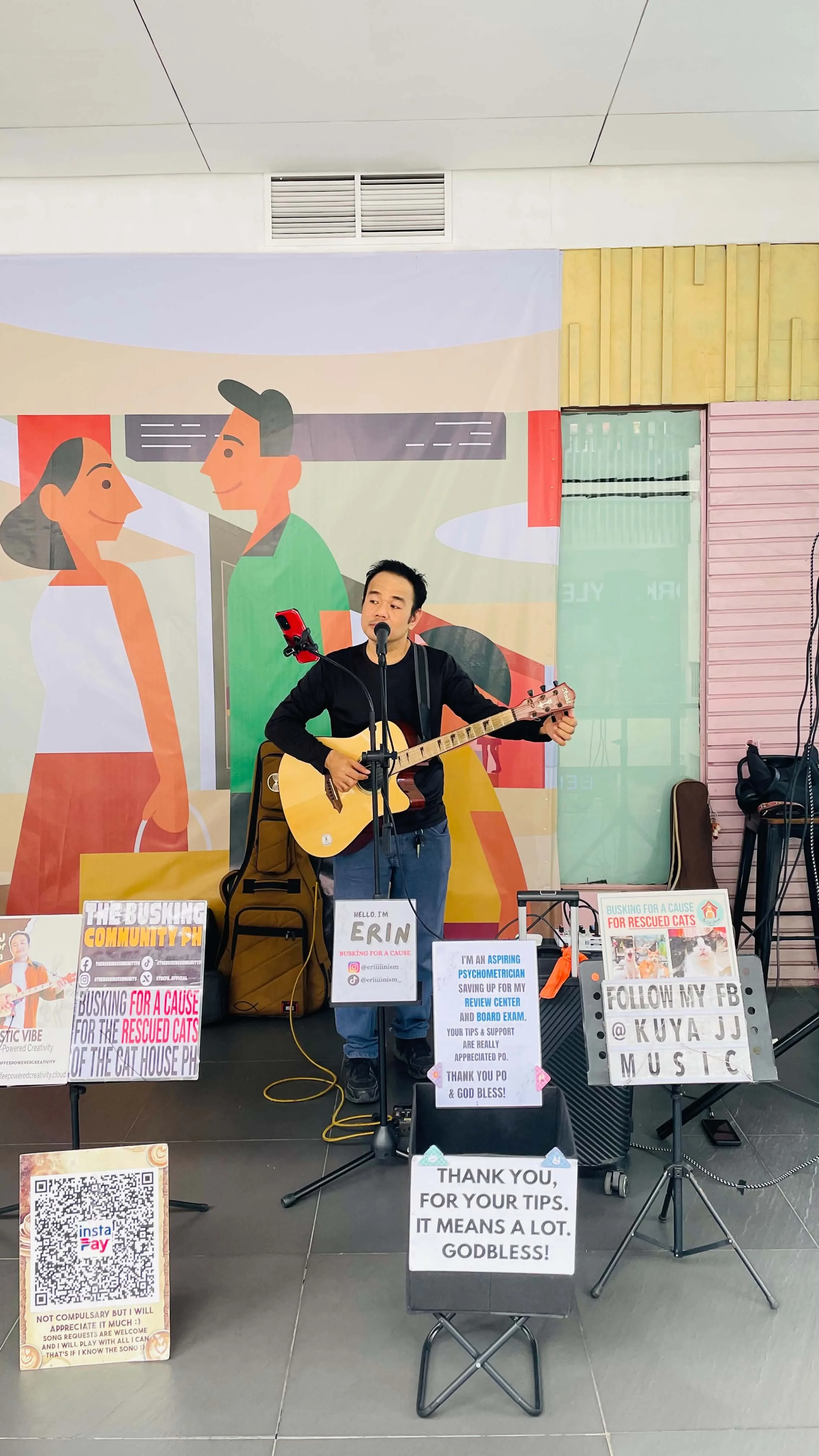 A young man playing guitar and singing into a microphone at a charity event with various signs and posters in the foreground.