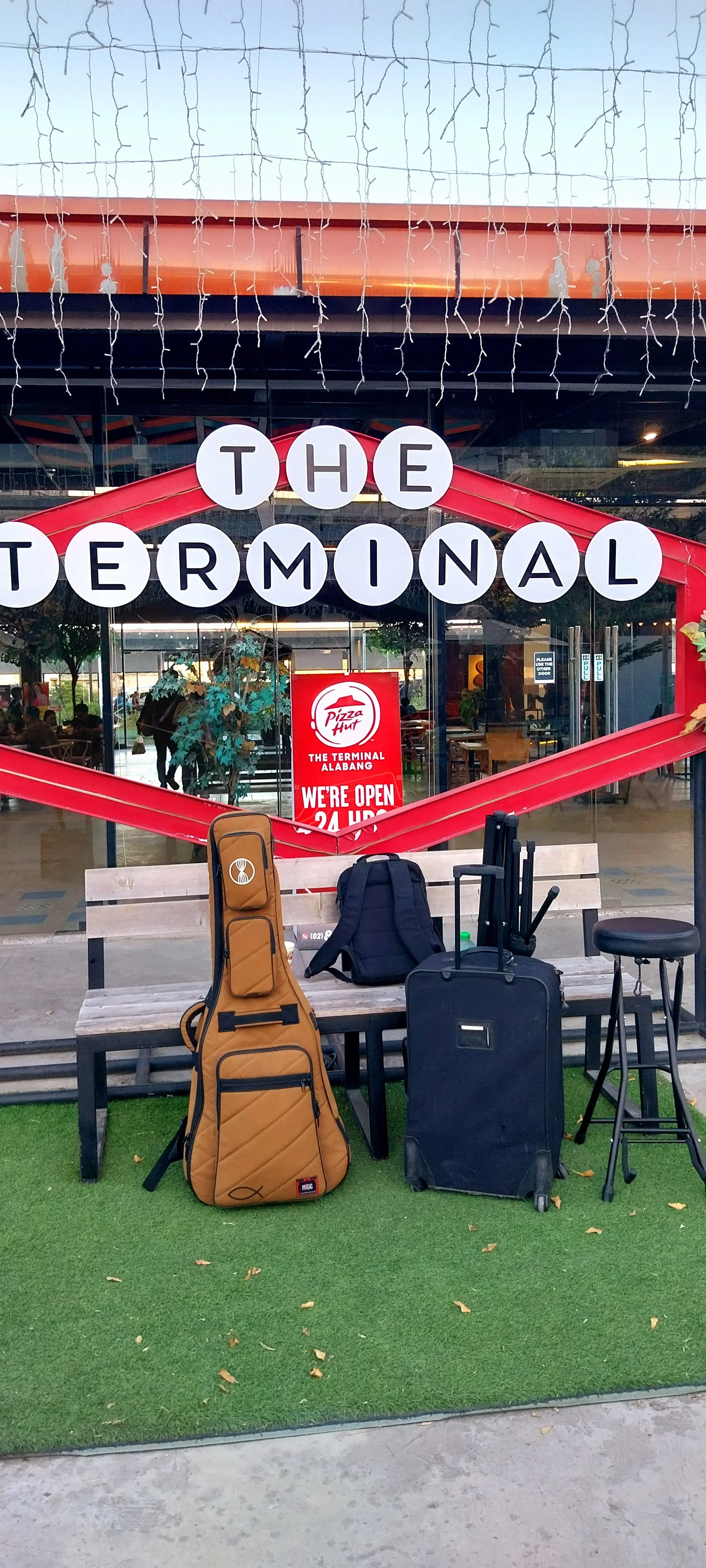 Storefront with sign reading 'The Terminal' and 'Pizza Hut,' luggage and a guitar case on a bench outside, with a green artificial grass mat and a stool in front.