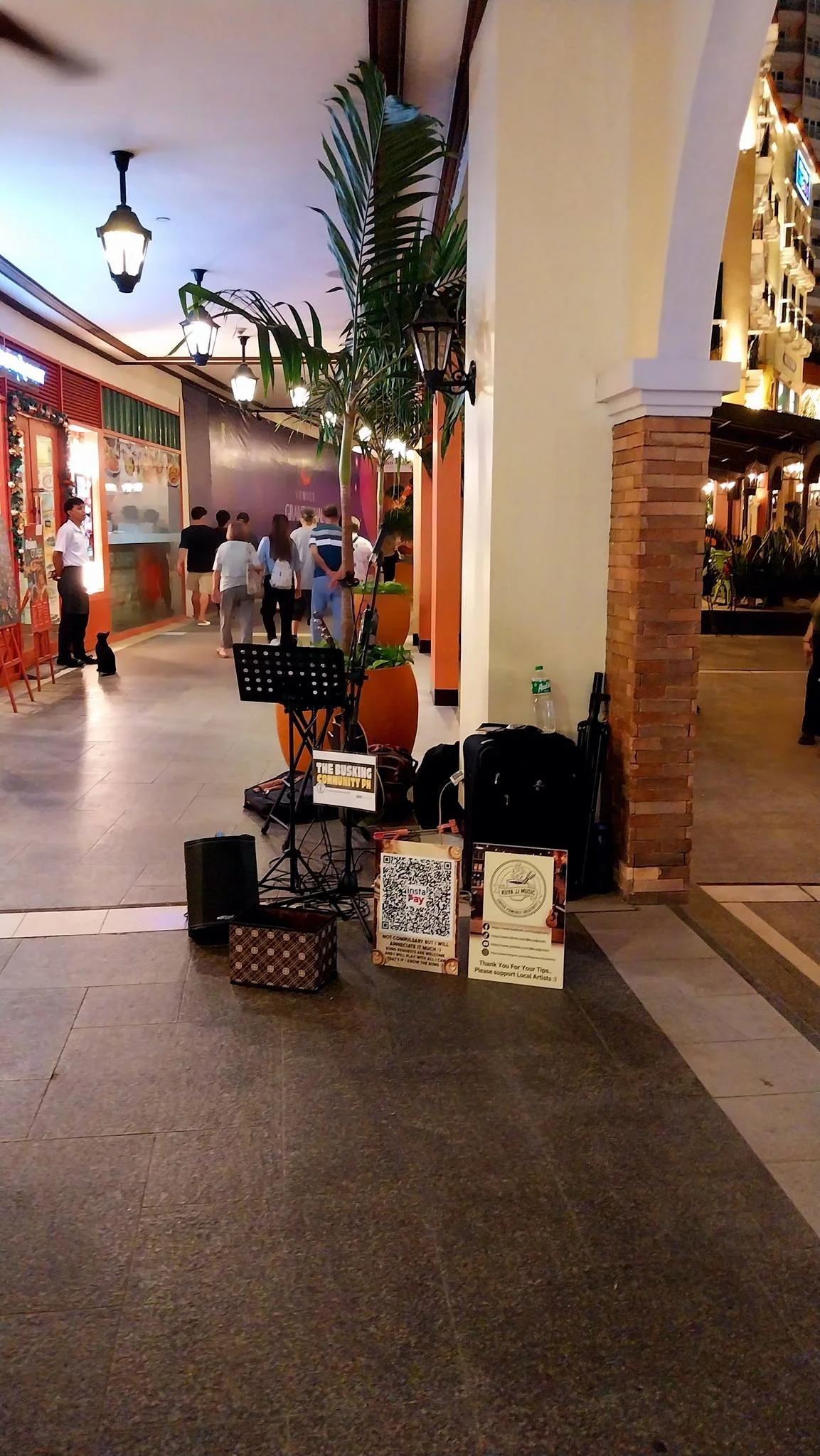 A street performer setup with a microphone stand, speaker, and donation signs in an indoor shopping mall corridor, with people walking in the background.