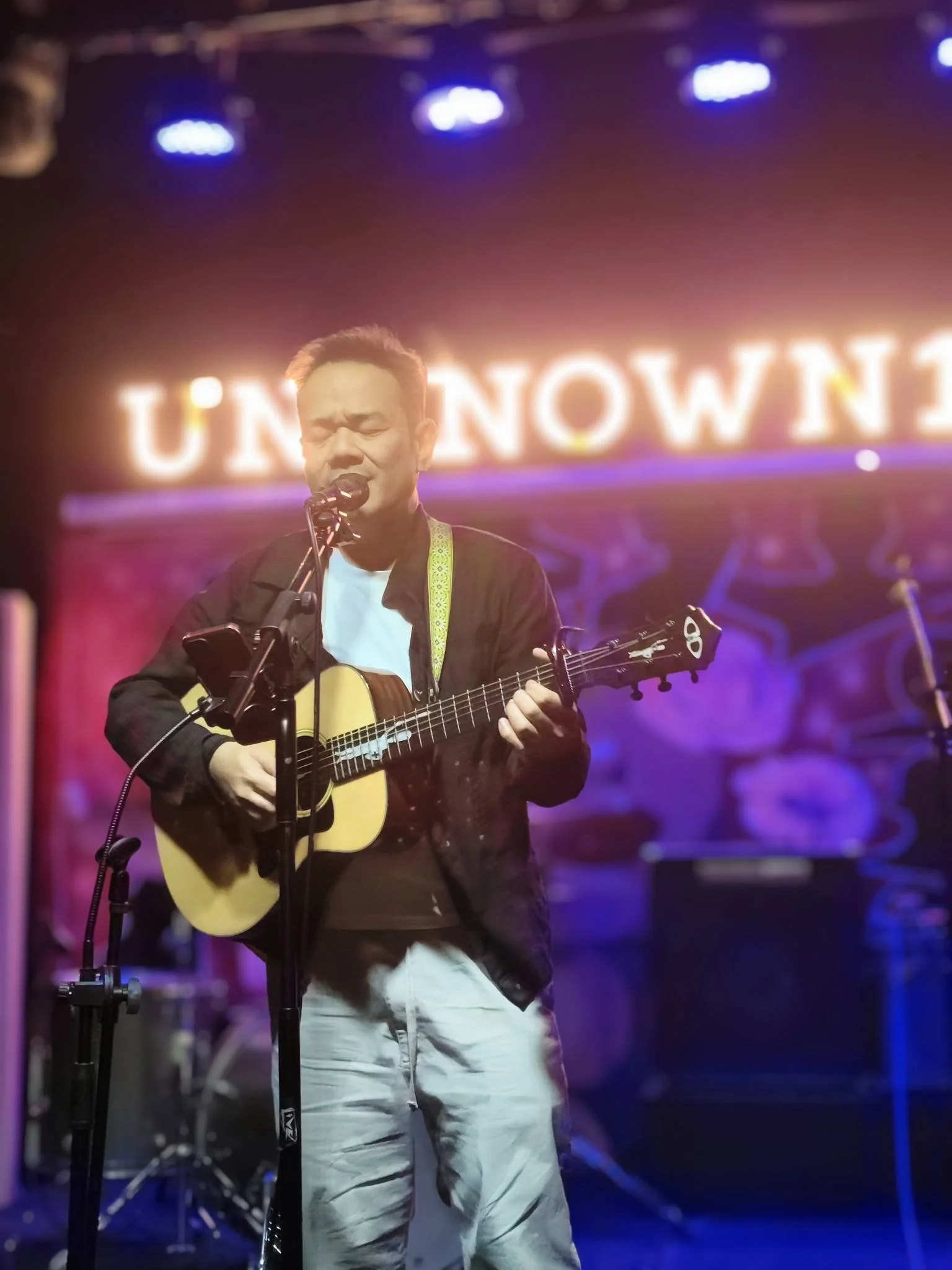 Man singing and playing an acoustic guitar on stage with a neon 'UNKNOWN' sign in the background.