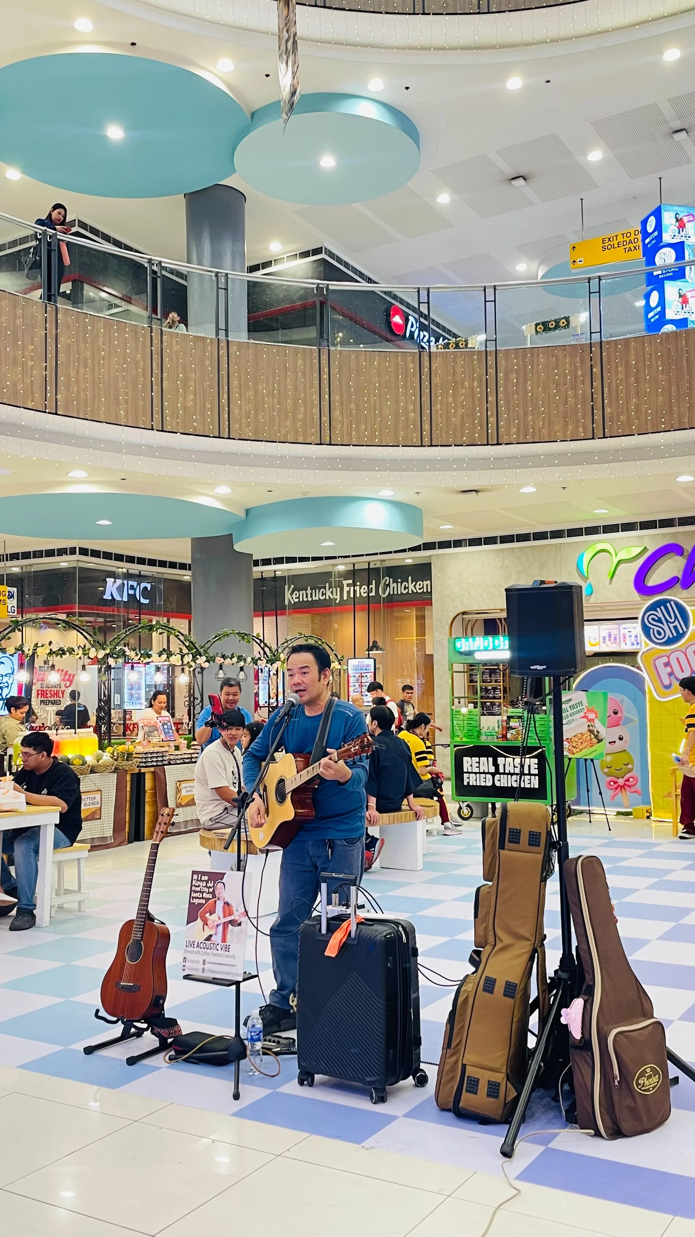 A man playing guitar and singing into a microphone in a shopping mall food court. There are shopping bags, guitars, and musical equipment around him. People are sitting and standing in the background, and there are various stores like Kentucky Fried 