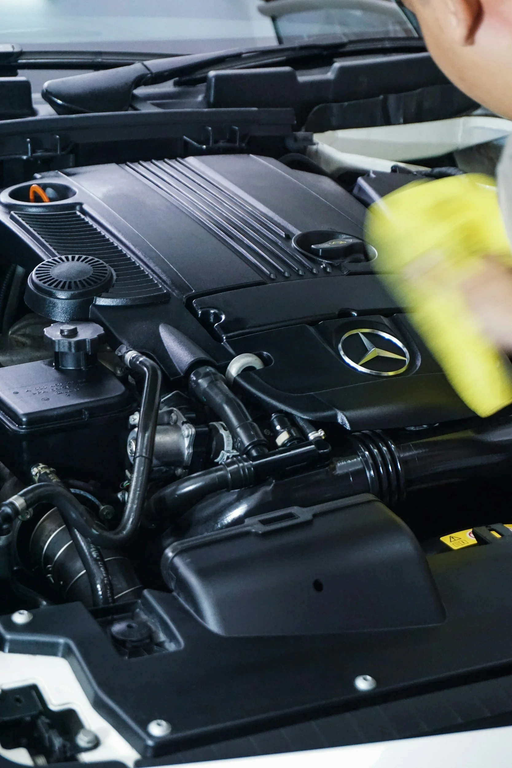 Close-up of a car engine with a person cleaning it using a yellow cloth, Mercedes-Benz logo visible on engine cover.