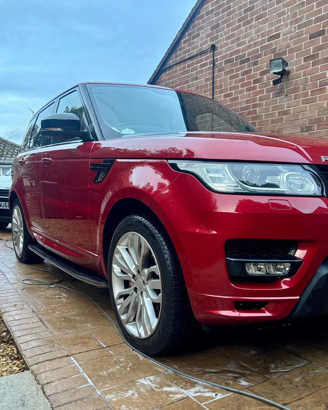 Red Range Rover parked on a wet driveway, with a brick wall and outdoor light fixture in the background.