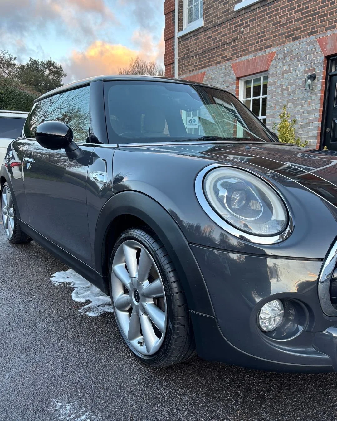 Black Mini Cooper parked on driveway with soap suds underneath, in front of a brick house, during sunset with partly cloudy sky.