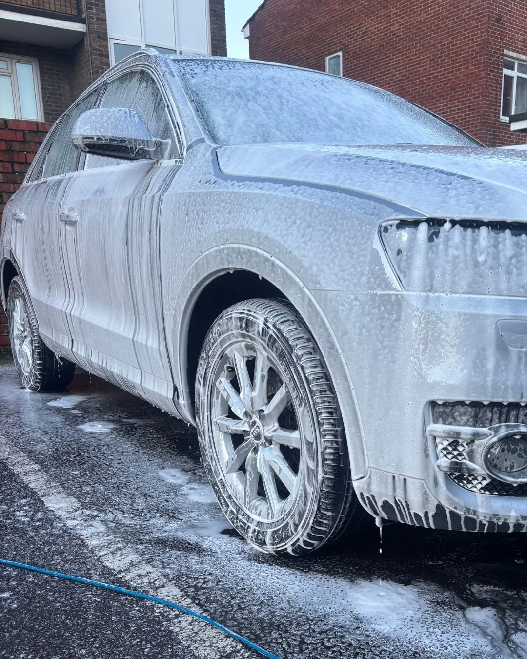 A silver car being washed with soap and foam, parked on a wet driveway, with foam covering parts of the car and surrounding ground.