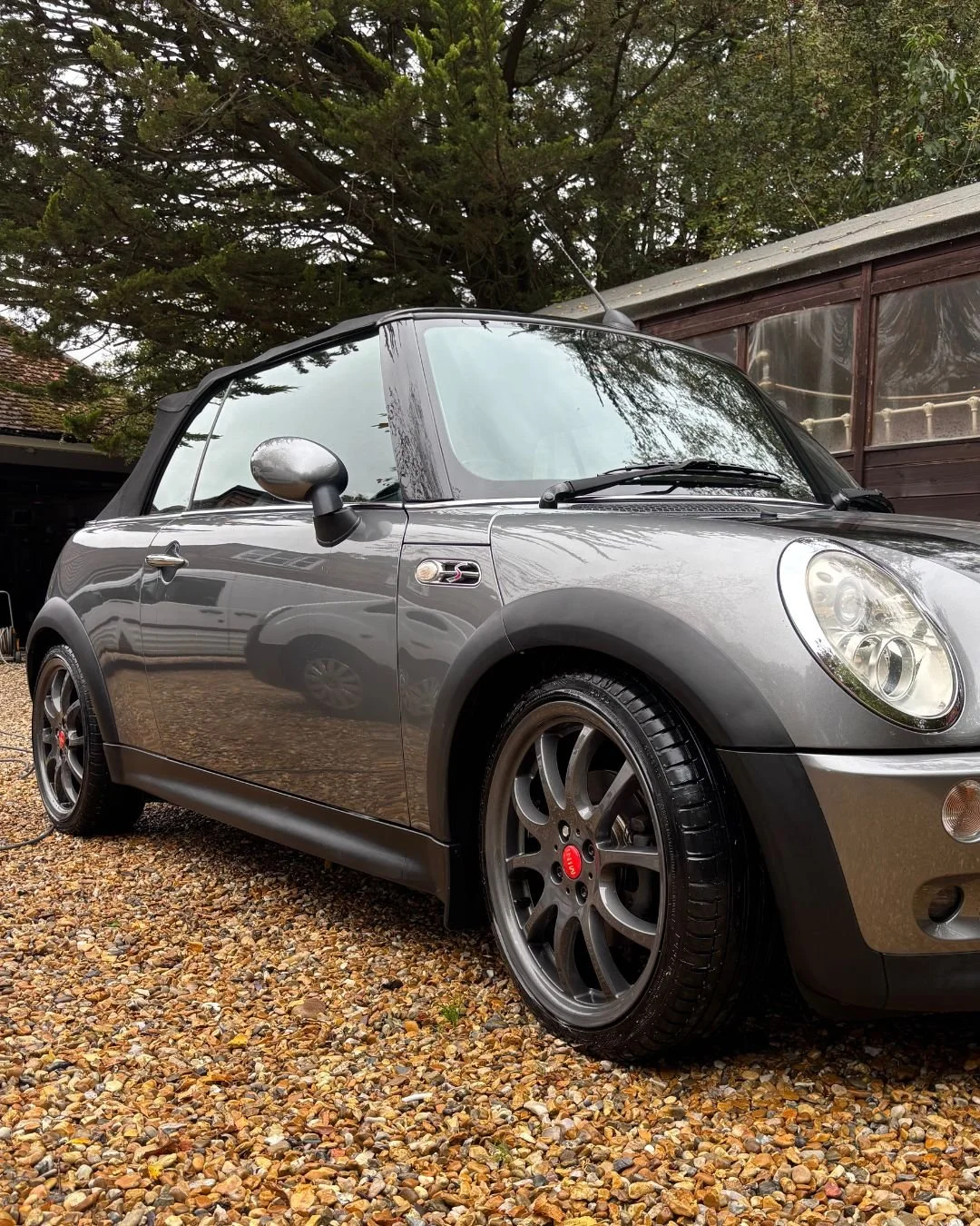 A gray Mini Cooper convertible parked on a gravel driveway near a wooden shed, with trees in the background.