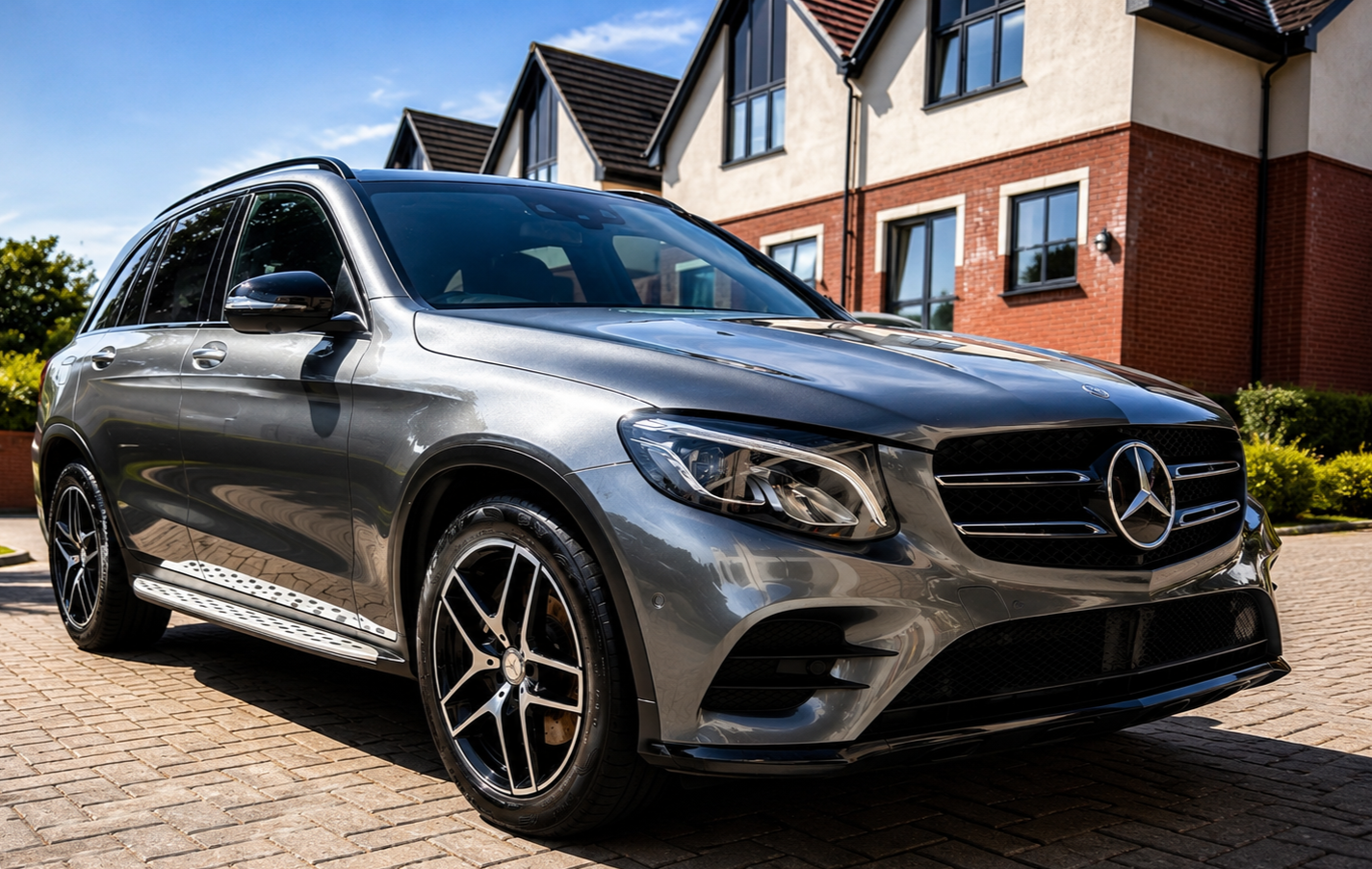 A silver Mercedes-Benz SUV parked on a brick driveway in front of a residential house with red brick and beige siding.