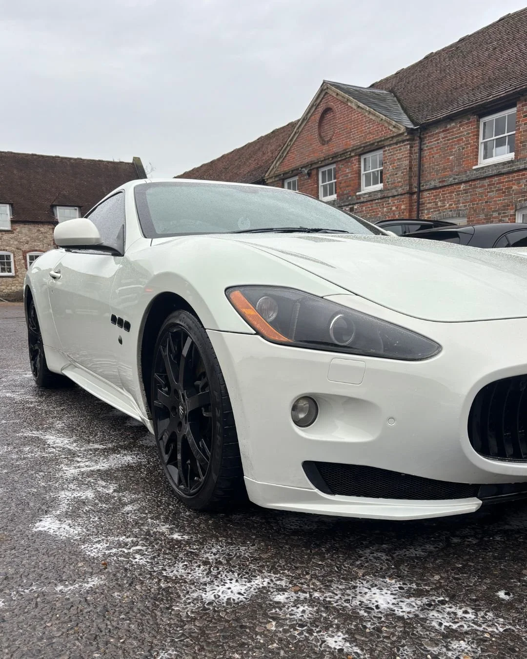 White sports car parked on wet pavement in front of brick house with red tiled roof, overcast sky.