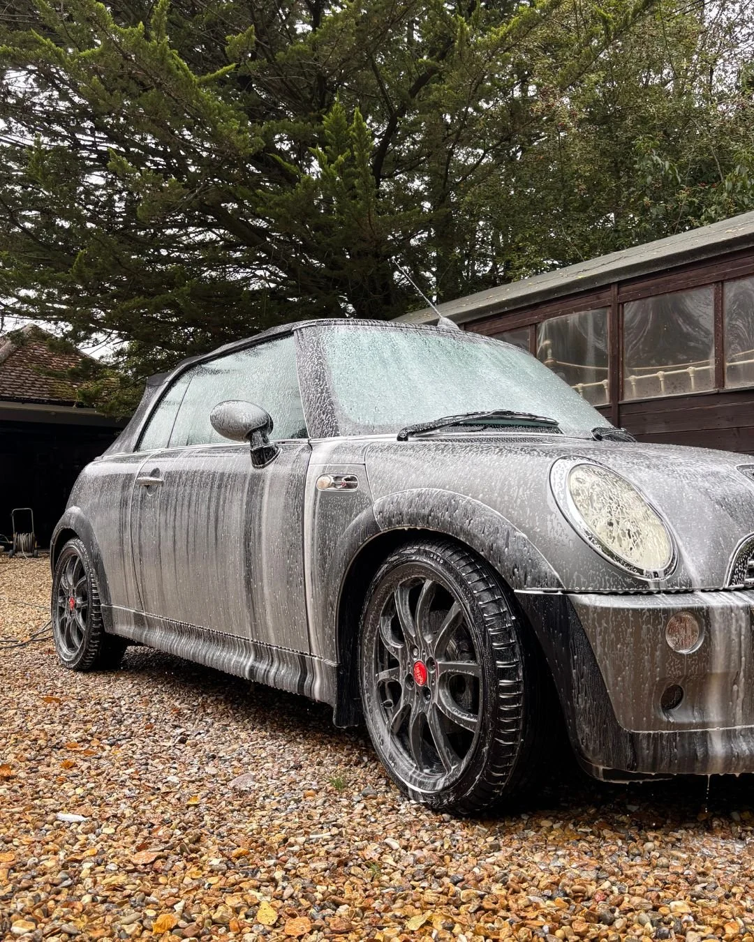 A small car covered in foam and soap, during a car wash, parked on a gravel driveway near a wooden shed with trees in the background.