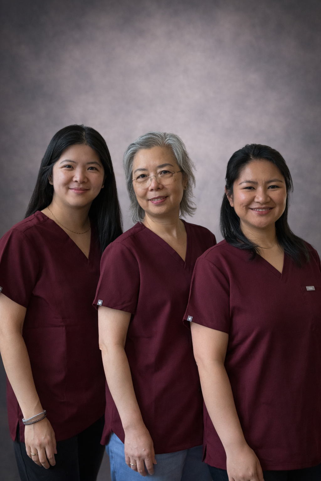 Three women wearing matching maroon medical scrubs standing side by side, smiling against a plain, gradient background.