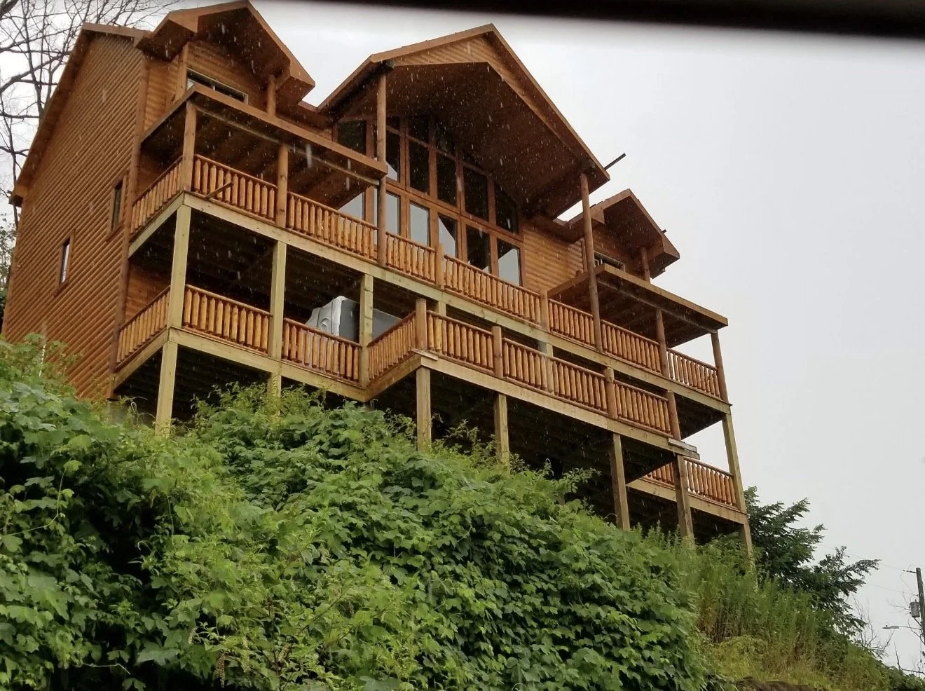 A multi-level log home with wide porches, balconies, and a sloped roof nestled into a hillside of dense green foliage. Every level includes porches finished with handcrafted custom log railing, safety, and an authentic Smoky Mountain look.