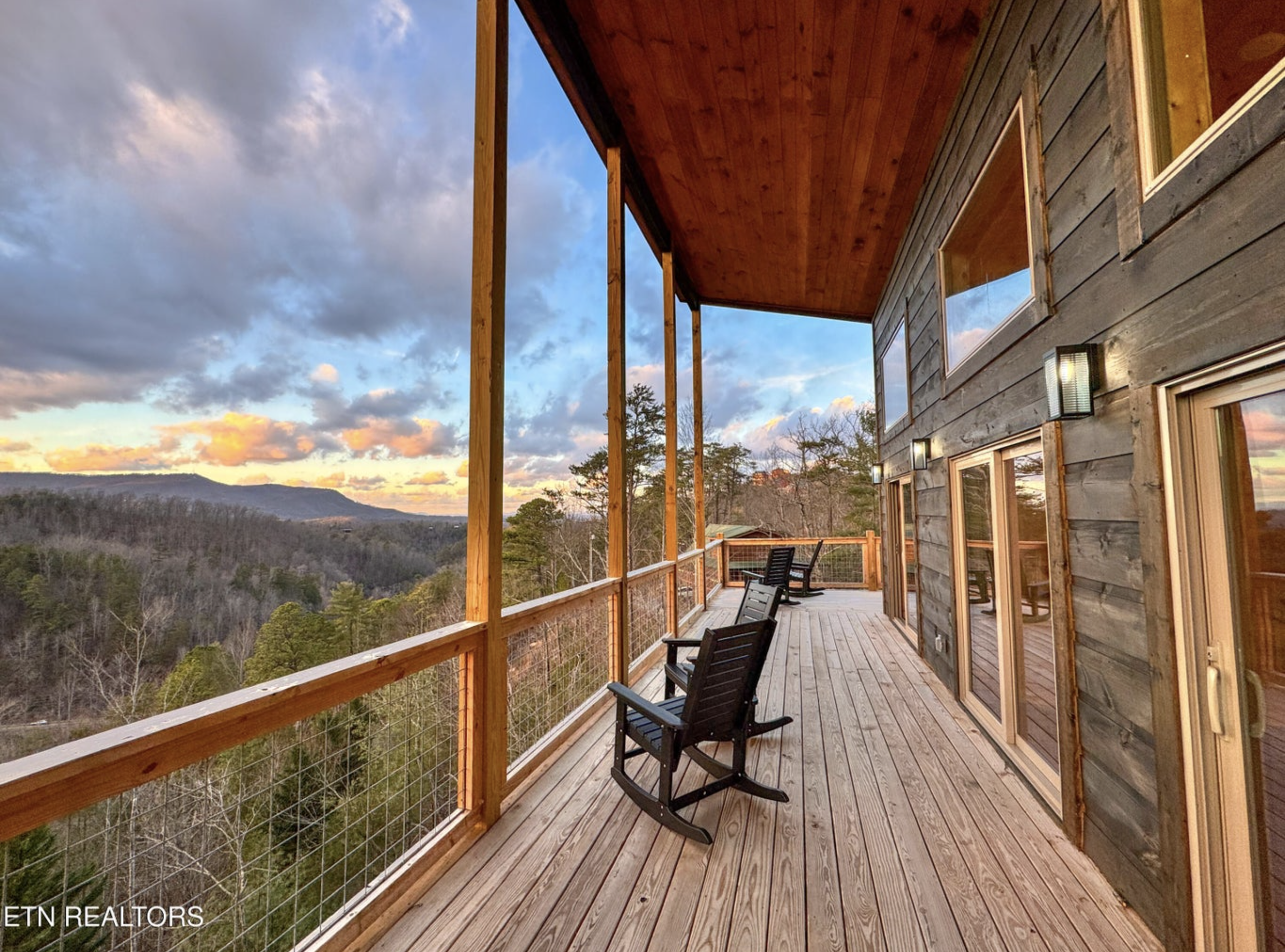 A spacious wooden balcony with rocking chairs overlooking a scenic mountain landscape at sunset.