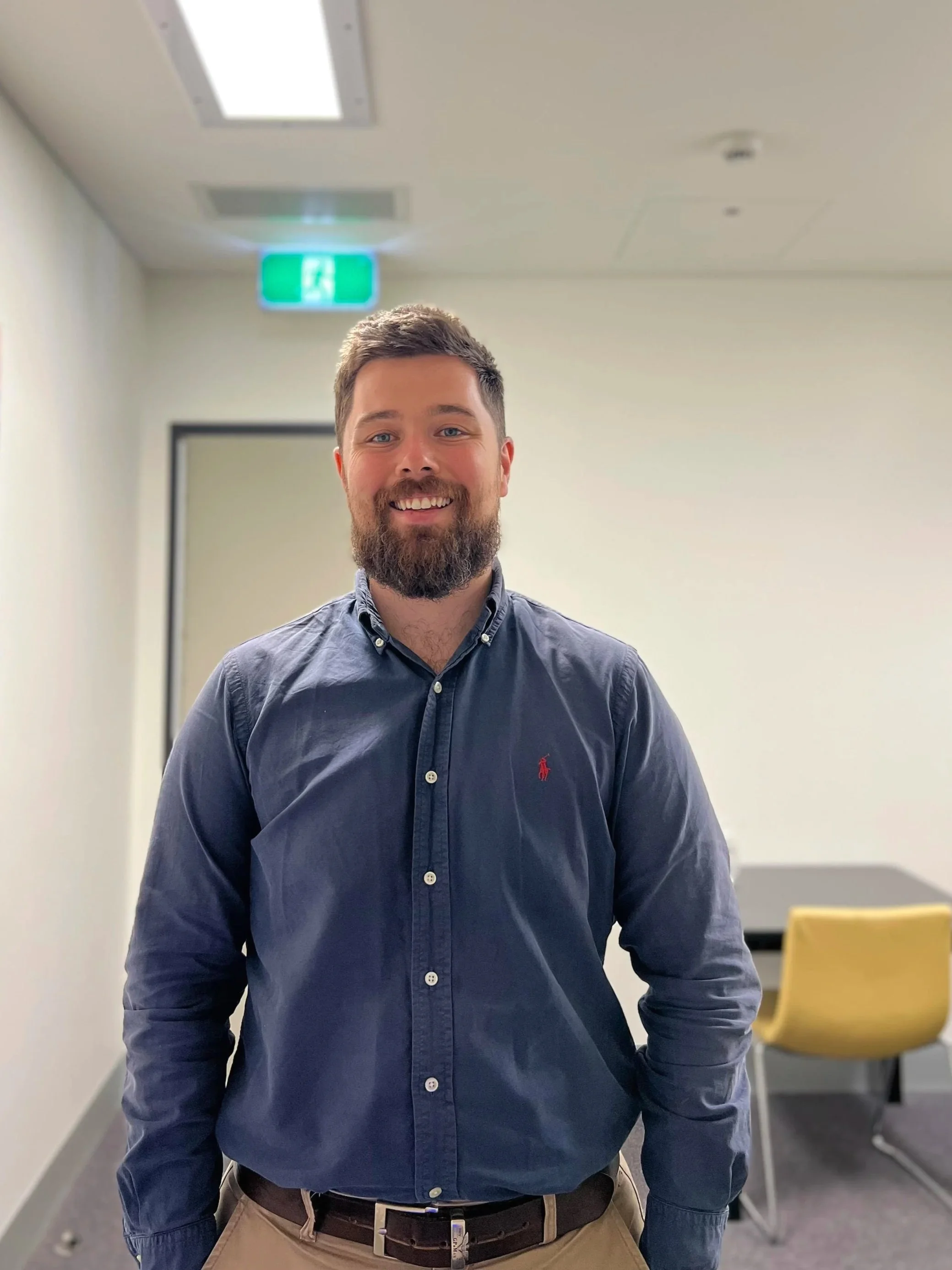 A smiling man with a beard and short hair, wearing a navy blue button-up shirt with a red Ralph Lauren logo, standing in a plain office corridor.