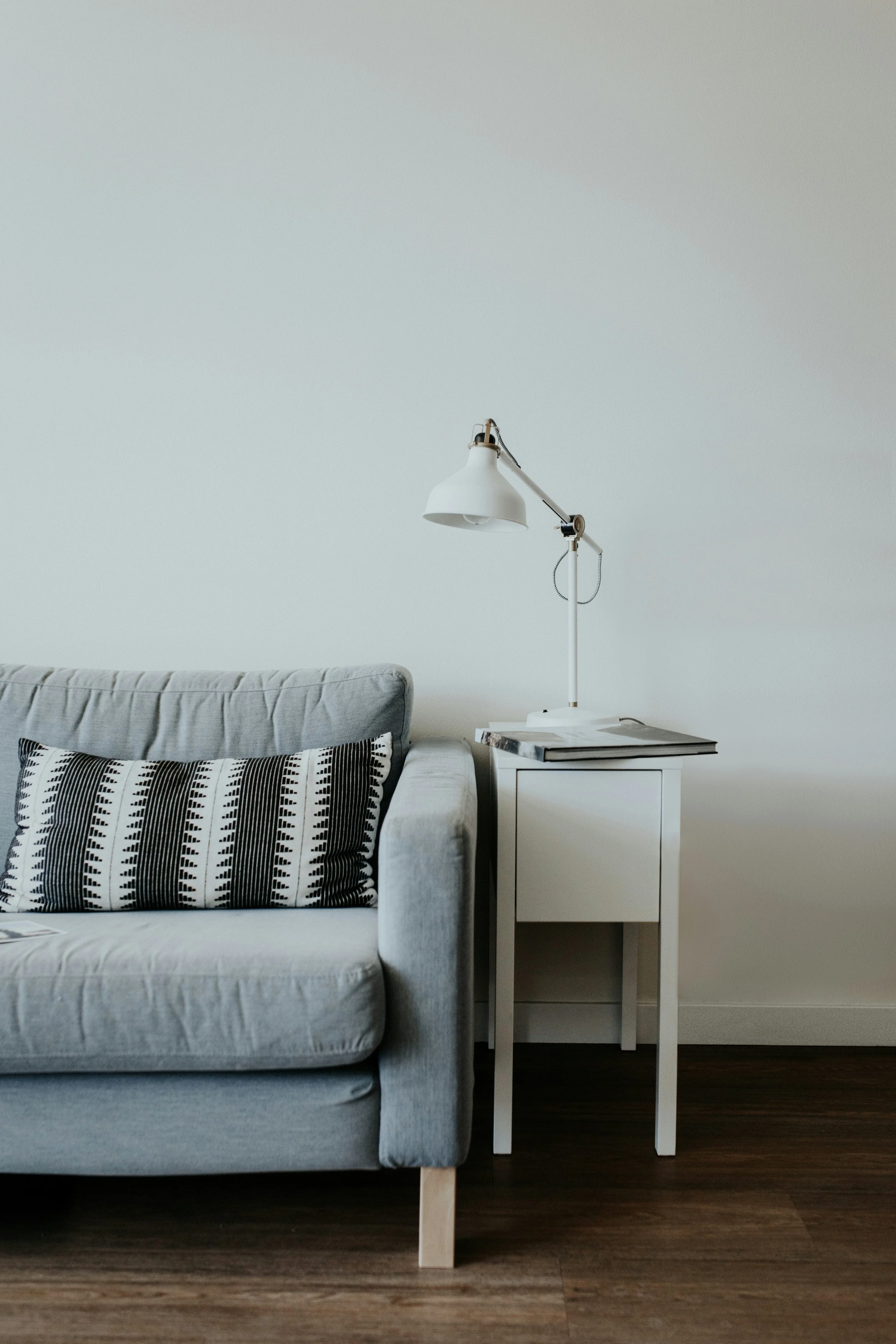 A living room corner with a light gray sofa, black and white patterned pillow, white side table, and a white adjustable desk lamp on the side table. Hardwood floor and plain off-white wall.