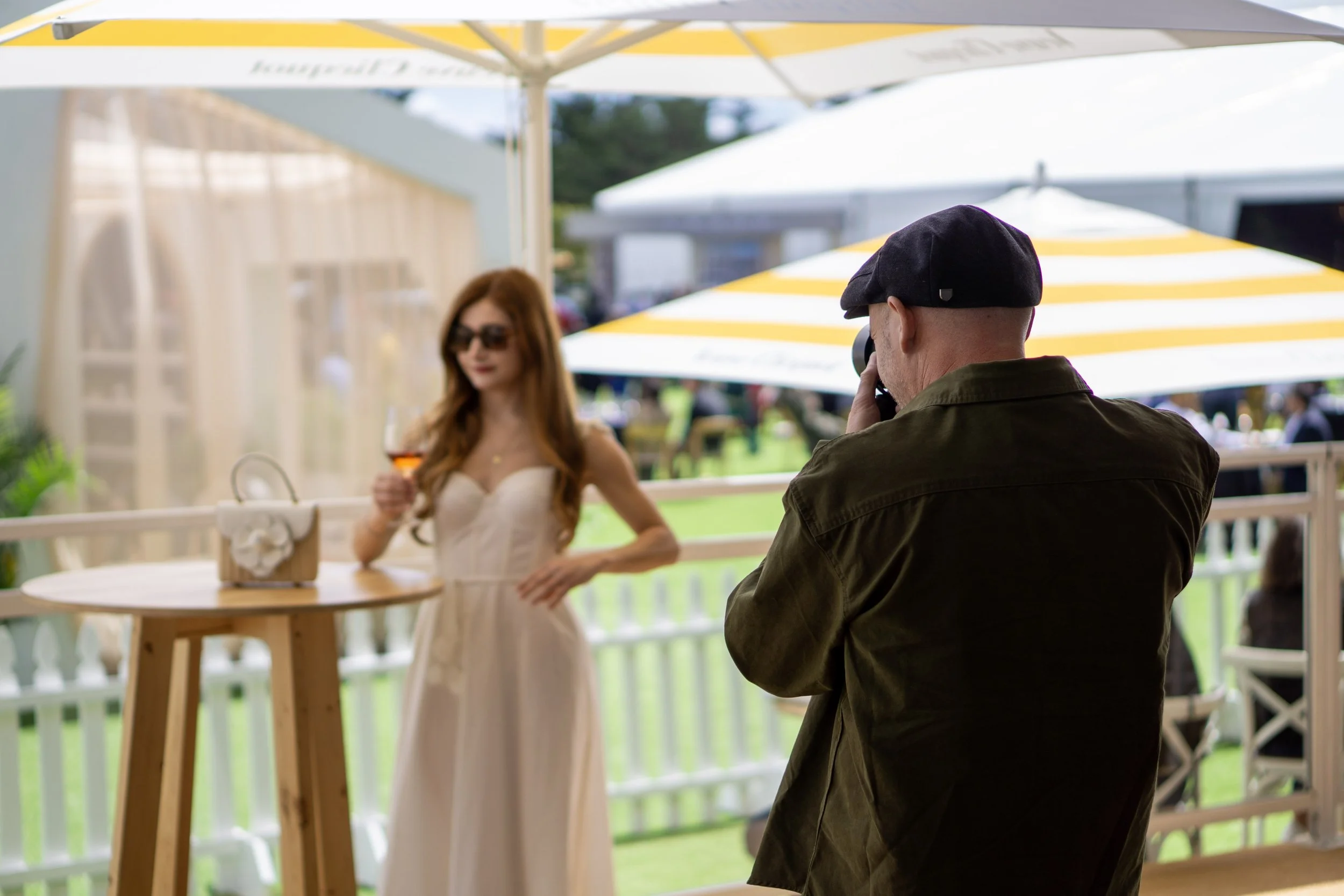 A man in a cap taking a photo of a woman in a beige dress holding a drink at an outdoor venue with yellow and white umbrellas.