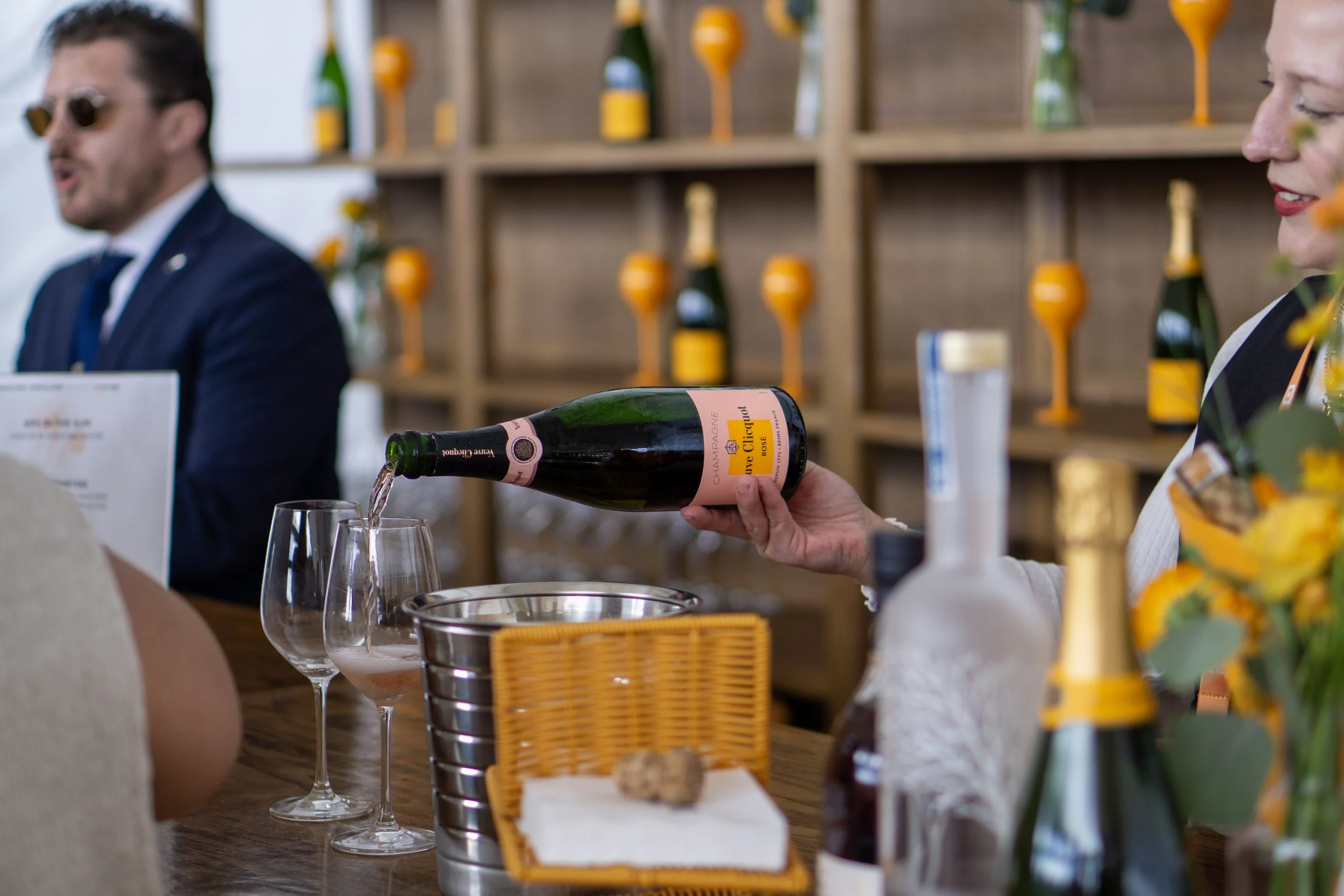 A person pours sparkling wine from a bottle into a glass at a bar or tasting event, with shelves of bottles and glasses in the background.