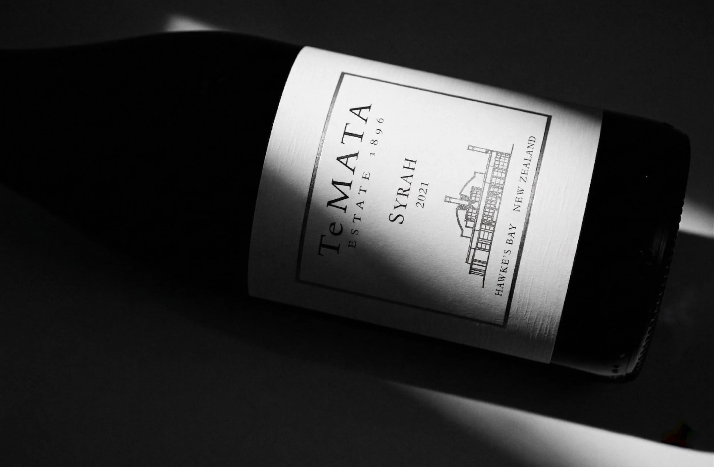 Black wine bottle lying on its side in a dark setting with a white label that reads 'Te Mata Estate 1896 Syriah 2021 Hawke's Bay New Zealand'.