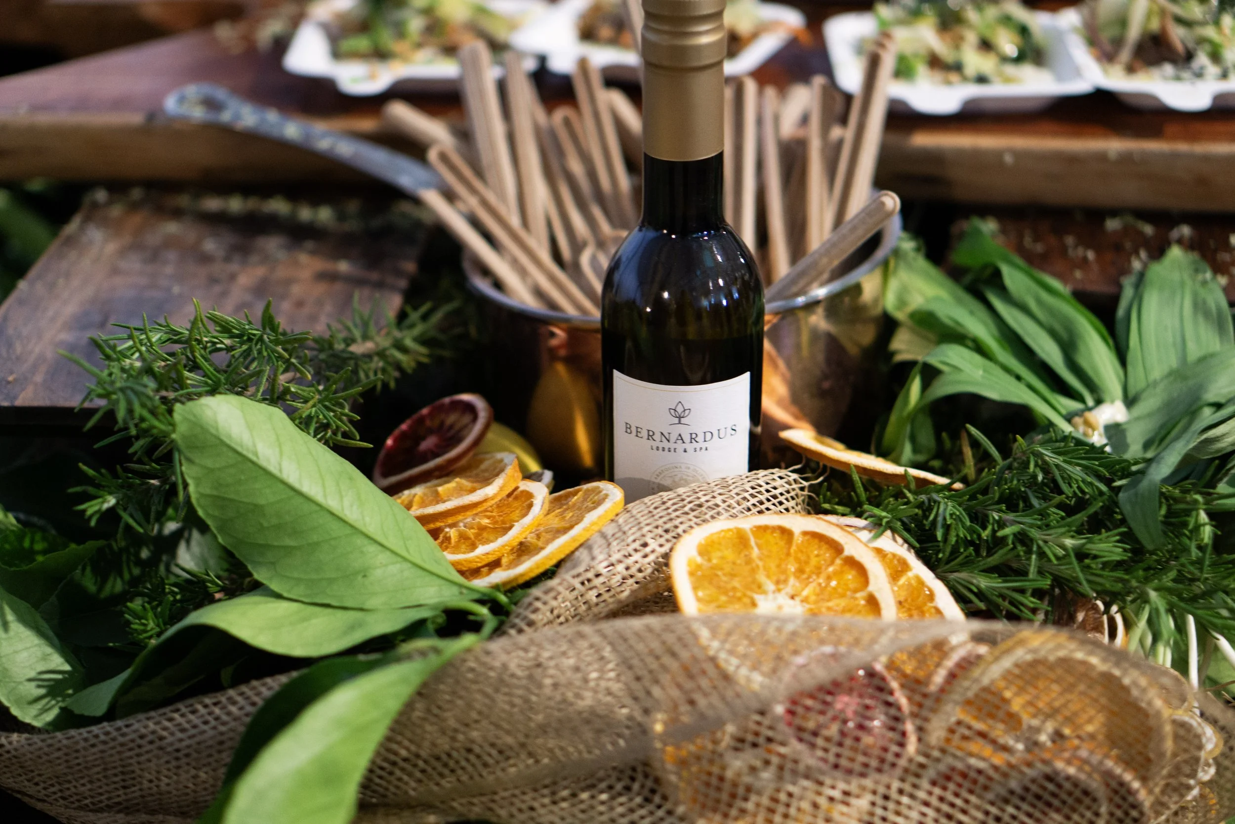 A wine bottle labeled 'Bernardus Lodge & Spa' surrounded by fresh herbs, citrus slices, and a woven cloth in a rustic setting.