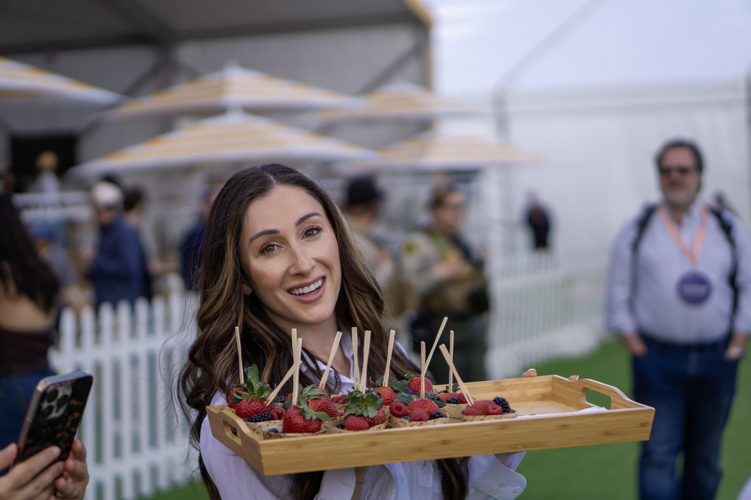 A woman holding a wooden tray filled with decorated cookies topped with fresh berries at an outdoor event with people and umbrellas in the background.