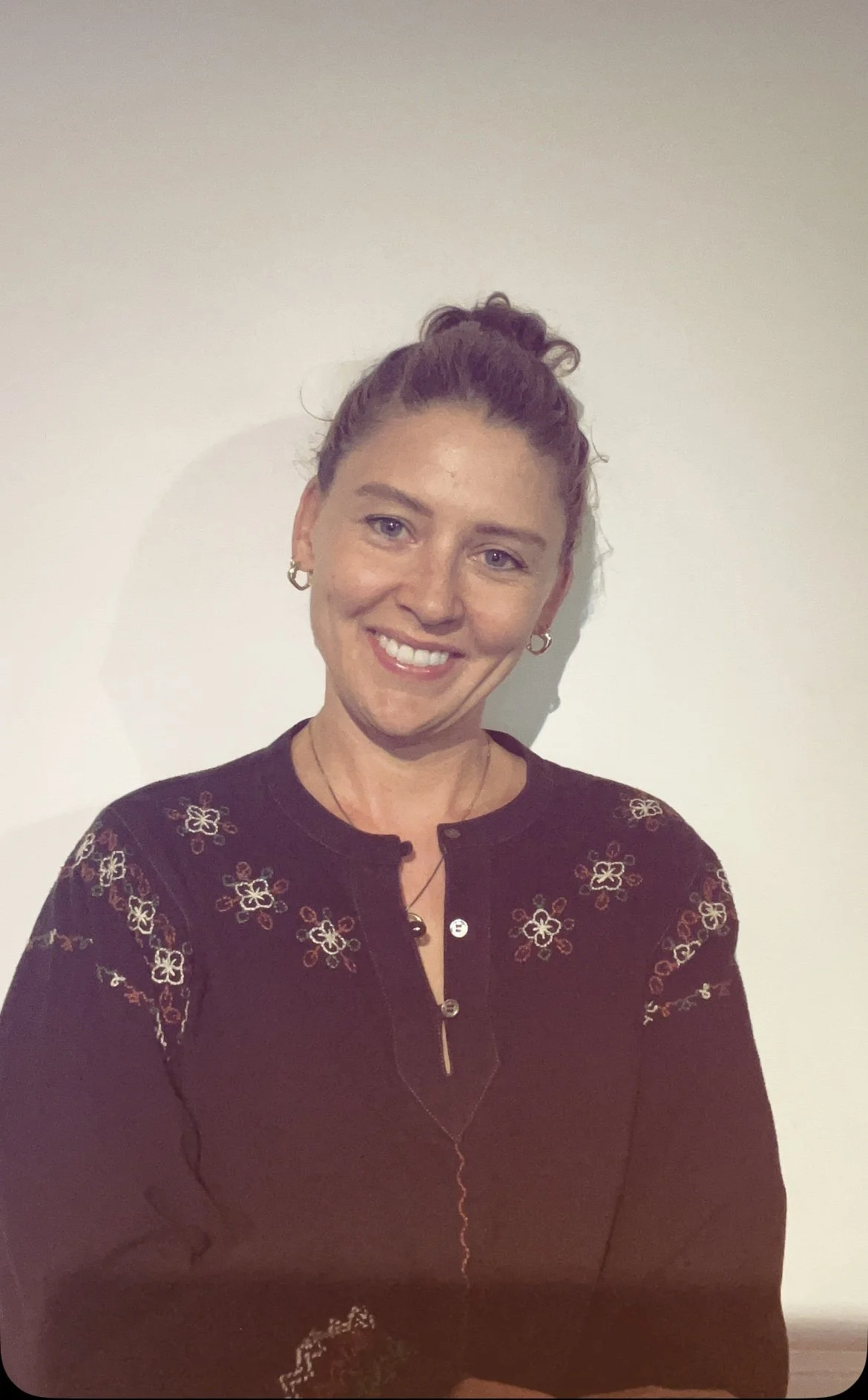 A woman with curly hair tied in a bun, smiling, wearing earrings and a dark blouse with floral embroidery, standing against a plain white wall.