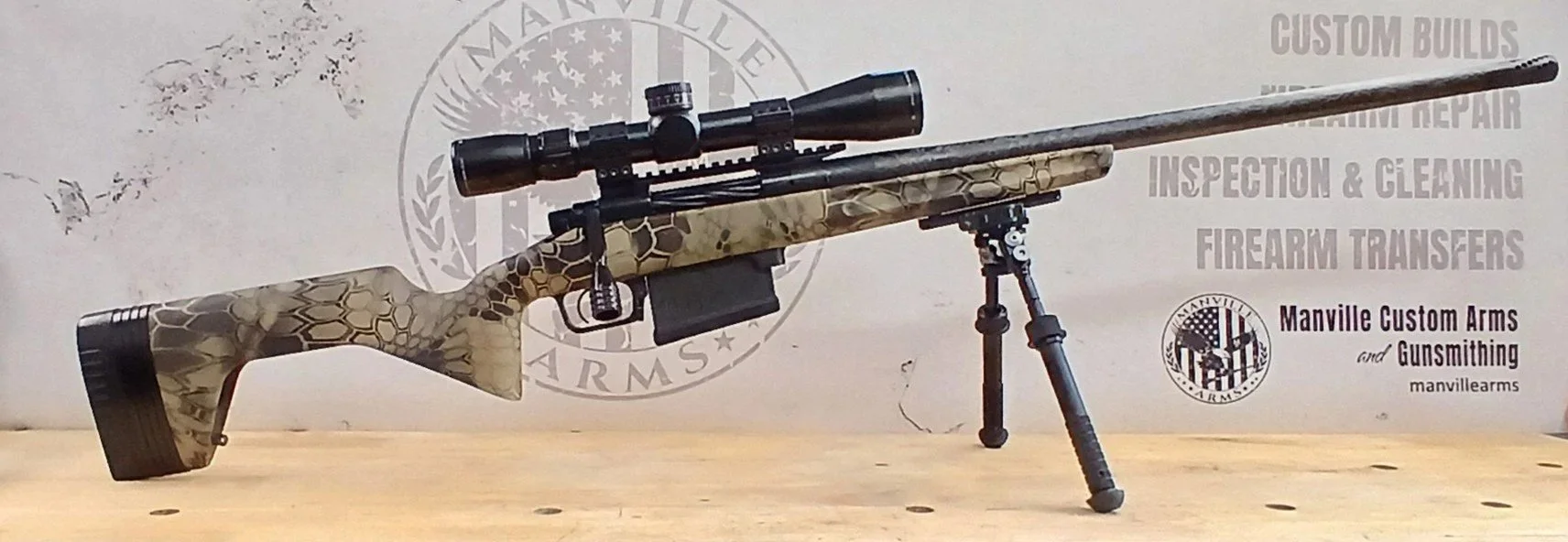 A scoped rifle with a camouflage pattern on the stock, mounted on a bipod, sitting on a wooden surface with a gray background featuring the Manville Arms logo and text about custom builds, firearm repair, inspection, cleaning, and transfers.