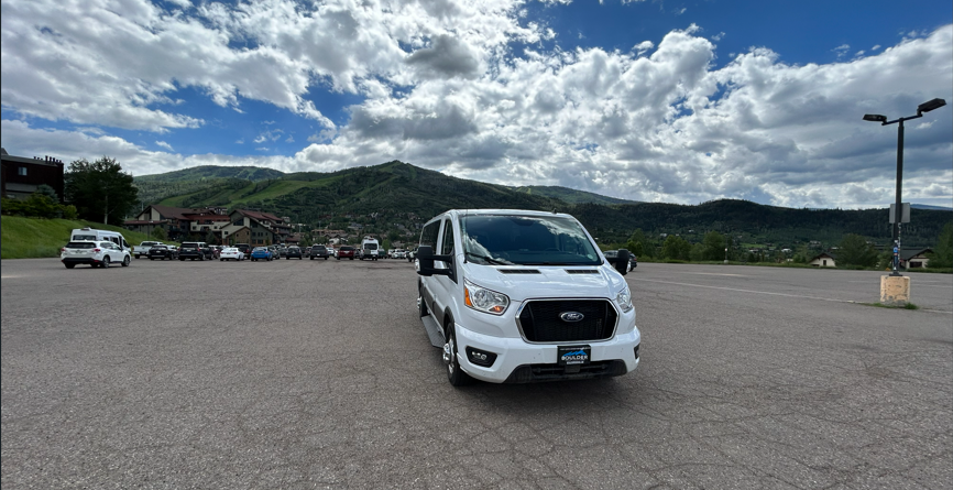 A white Ford Transit van parked in an empty parking lot with mountains and partly cloudy sky in the background.
