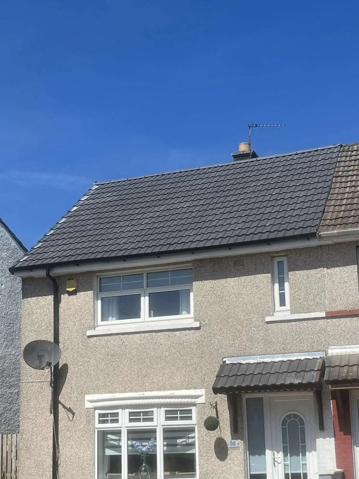 Front view of a two-story house with a pebbled exterior, white-framed windows, and a gray tiled roof. There is a satellite dish on the left side and an antenna on the roof under a clear blue sky.