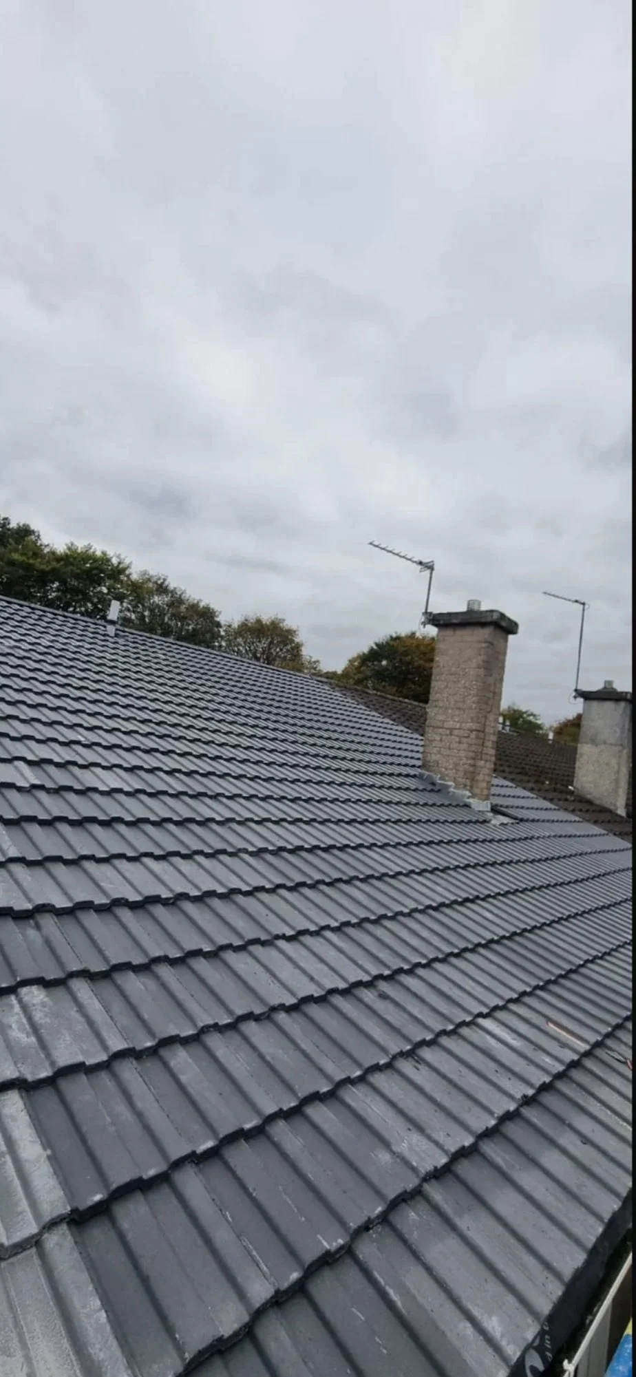 View of a gray tiled rooftop with two brick chimneys and television antennas under an overcast sky.