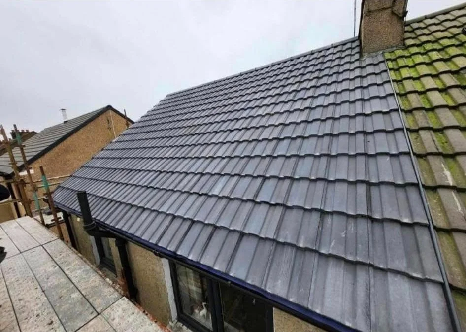 Close-up of a new black tiled roof on a house, with a neighboring roof visible on the right side. The house has a chimney and a window underneath the roof.