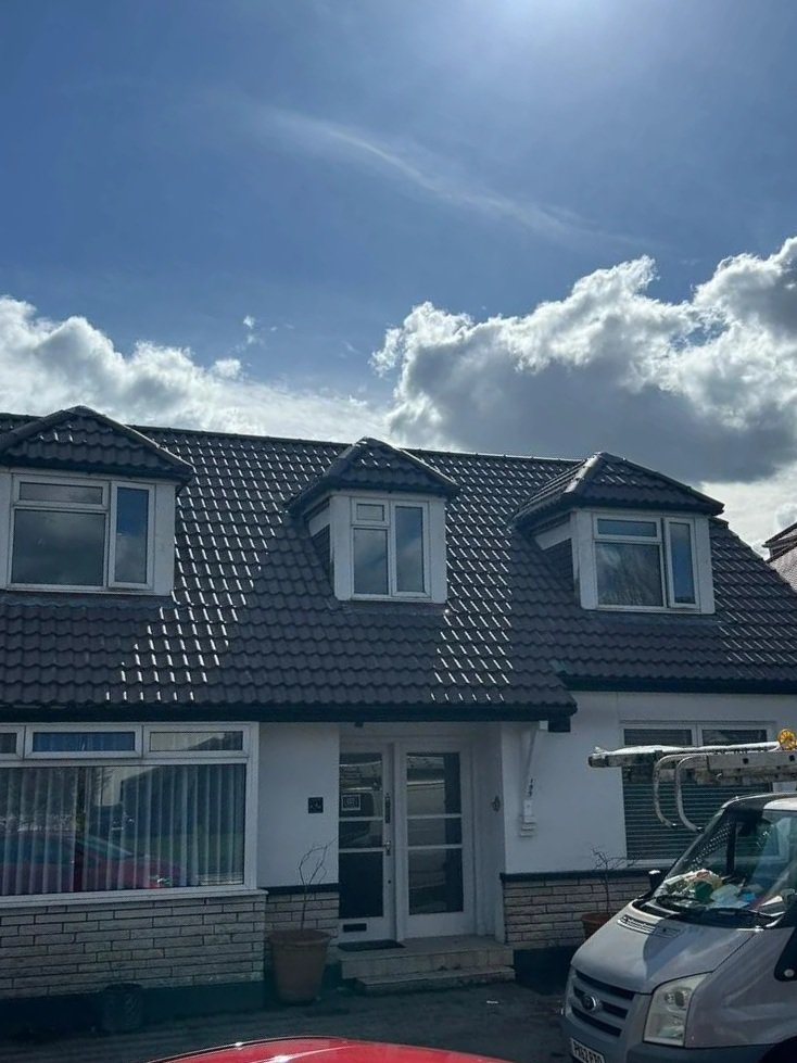 Front of a house with a black tiled roof, three bay windows, and a white door; a cloudy sky above and a white van parked nearby.