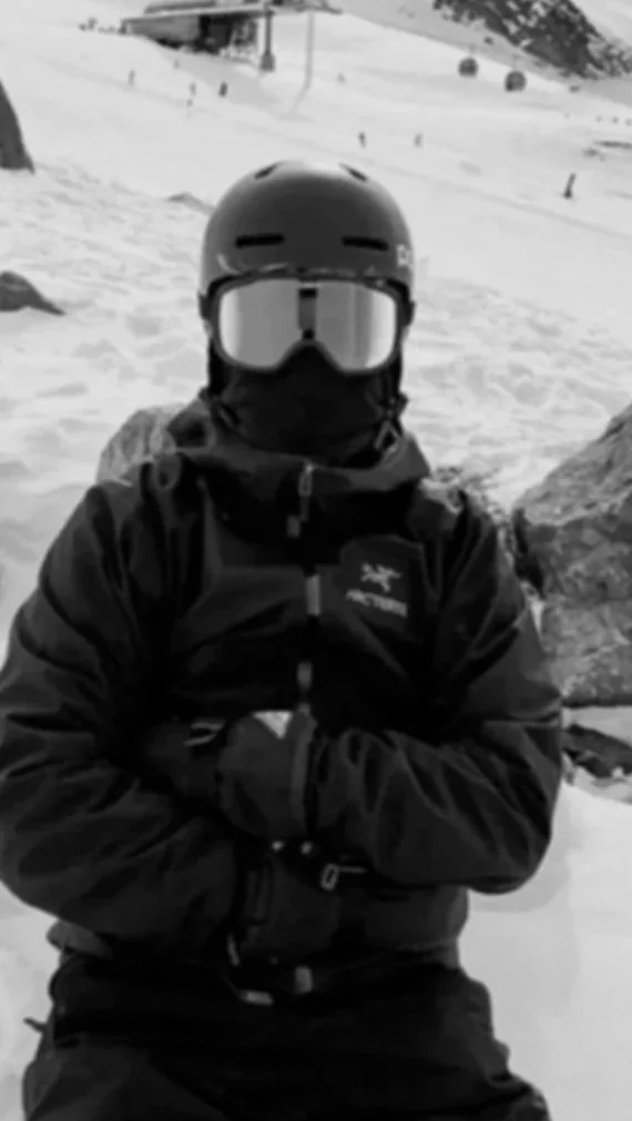 Person dressed in black winter gear, including a helmet, ski mask, and goggles, sitting in a snowy mountain landscape.