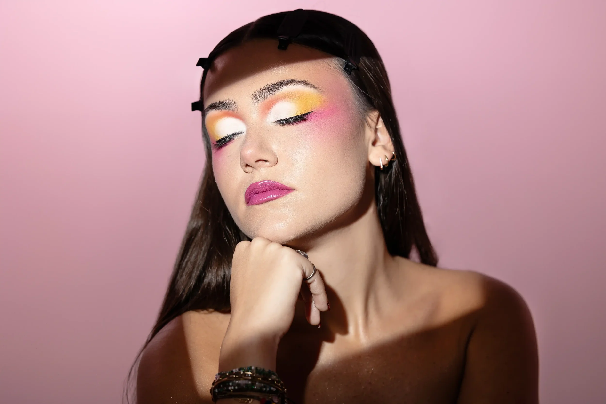 Jeune femme avec maquillage coloré, yeux fermés, peau claire, cheveux longs, portant plusieurs bracelets, avec un fond rose