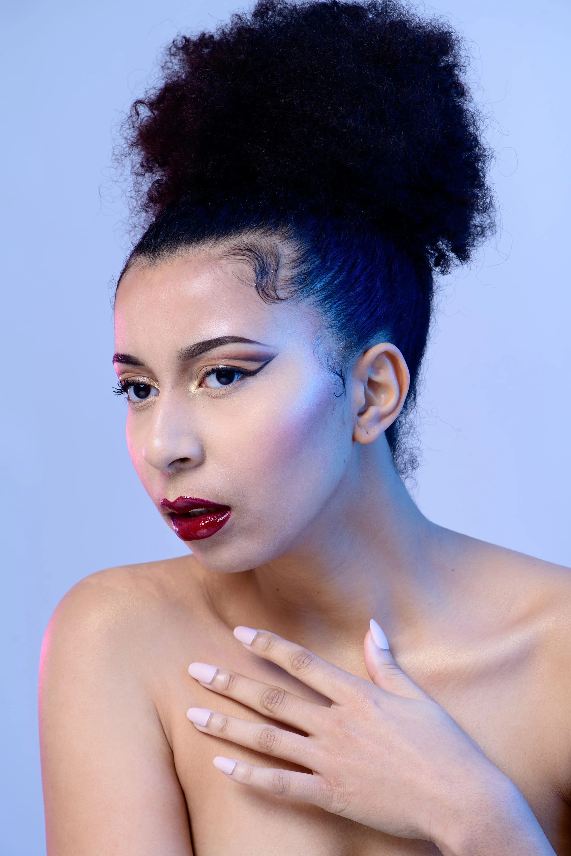 Un portrait d'une femme avec une coiffure volumineuse et bouclée en haut, maquillage audacieux avec œil de chat, lèvres rouges foncées, et une peau claire. Elle touche son épaule avec sa main gauche, le fond est de couleur bleue claire.