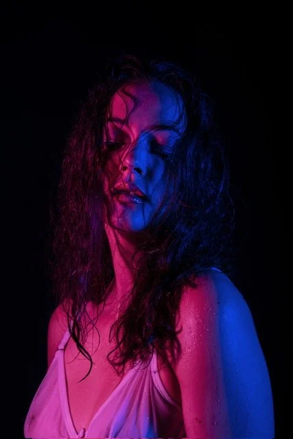 Une femme aux cheveux bouclés avec un maquillage coloré, éclairée en lumière rose et bleue sur fond noir.