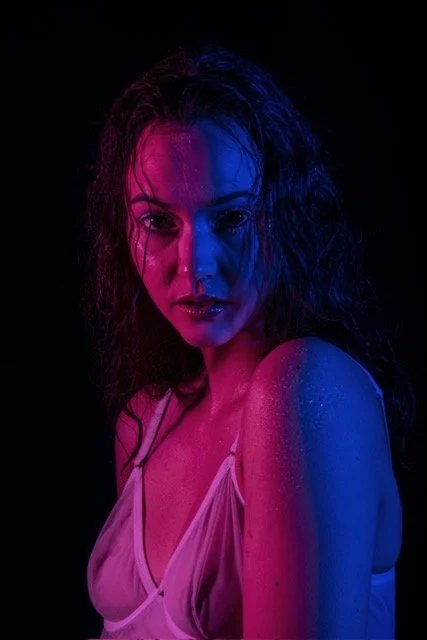 Portrait d'une femme avec des effets de lumière rose et bleue sur son visage et son corps, portant un chemisier léger, fond noir.