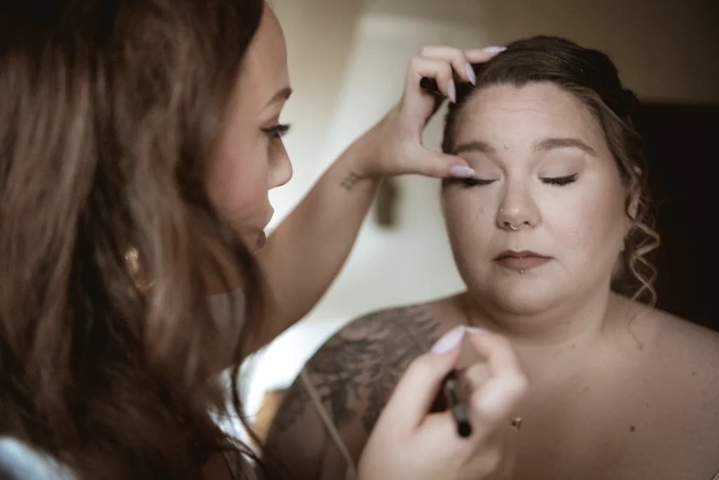 Une femme maquille une autre femme, qui a les yeux fermés, dans un intérieur