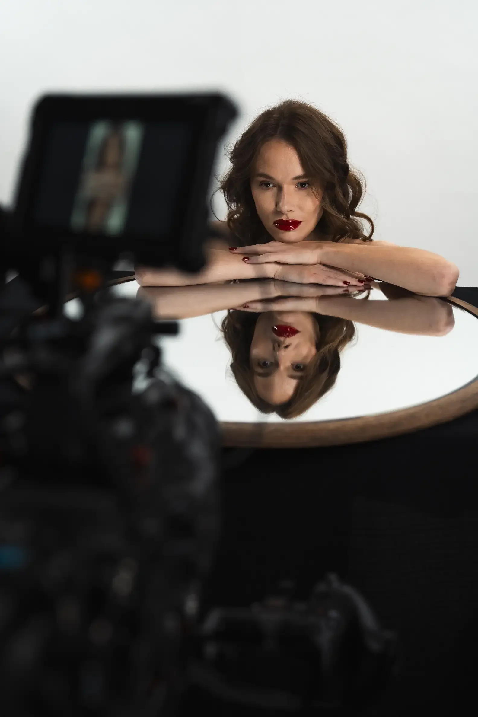 Une femme avec des cheveux bruns bouclés, portant du rouge à lèvres rouge foncé, regarde fixement l'objectif tout en posant avec ses bras reposant sur une surface réfléchissante, avec son reflet visible. Elle est en train de prendre une photo ou une vidéo d'elle-même avec un appareil photo numérique placé devant elle.