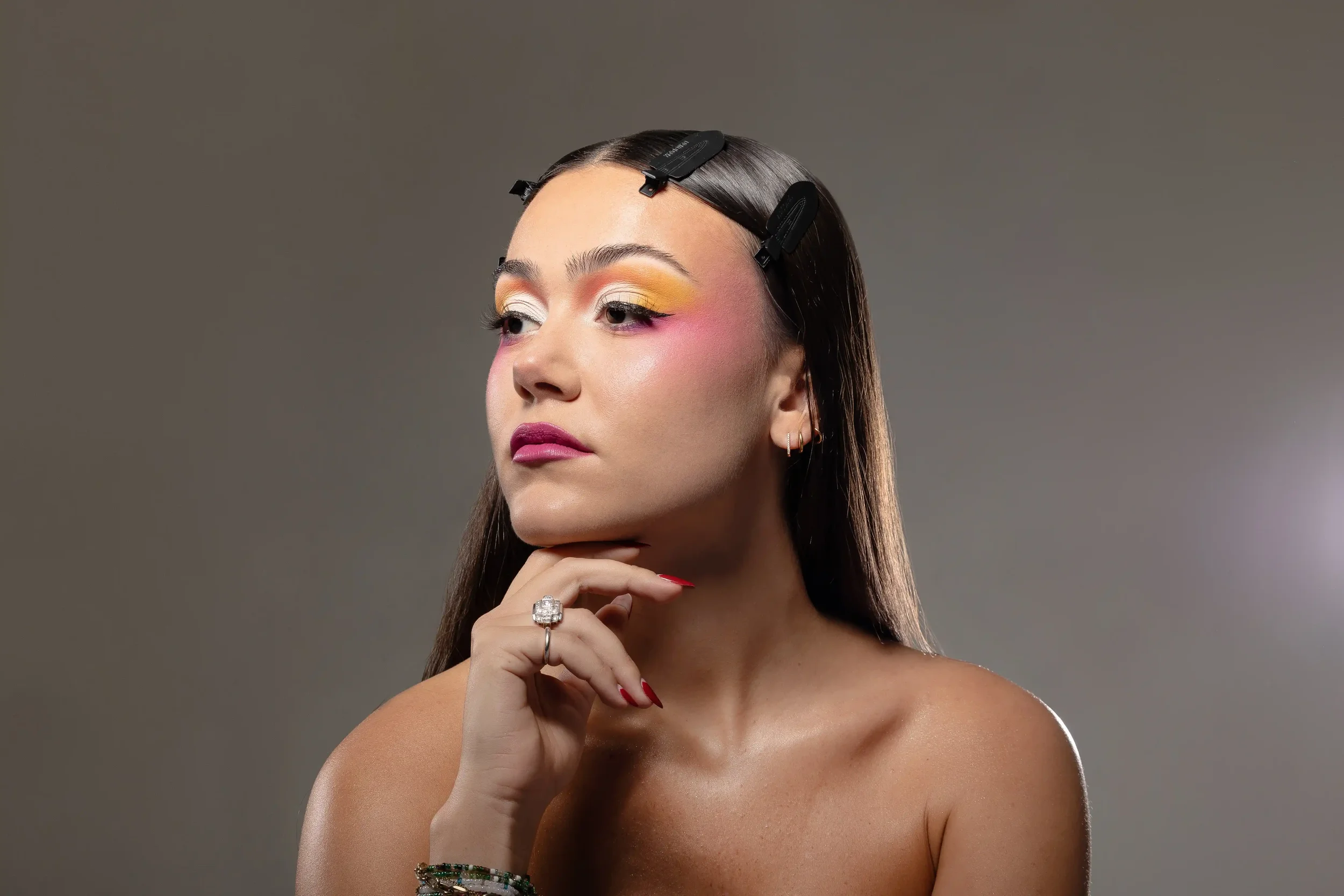 Une jeune femme avec des clips capillaires dans les cheveux, maquillage coloré, portant une bague et des bracelets, posant sur un fond gris.