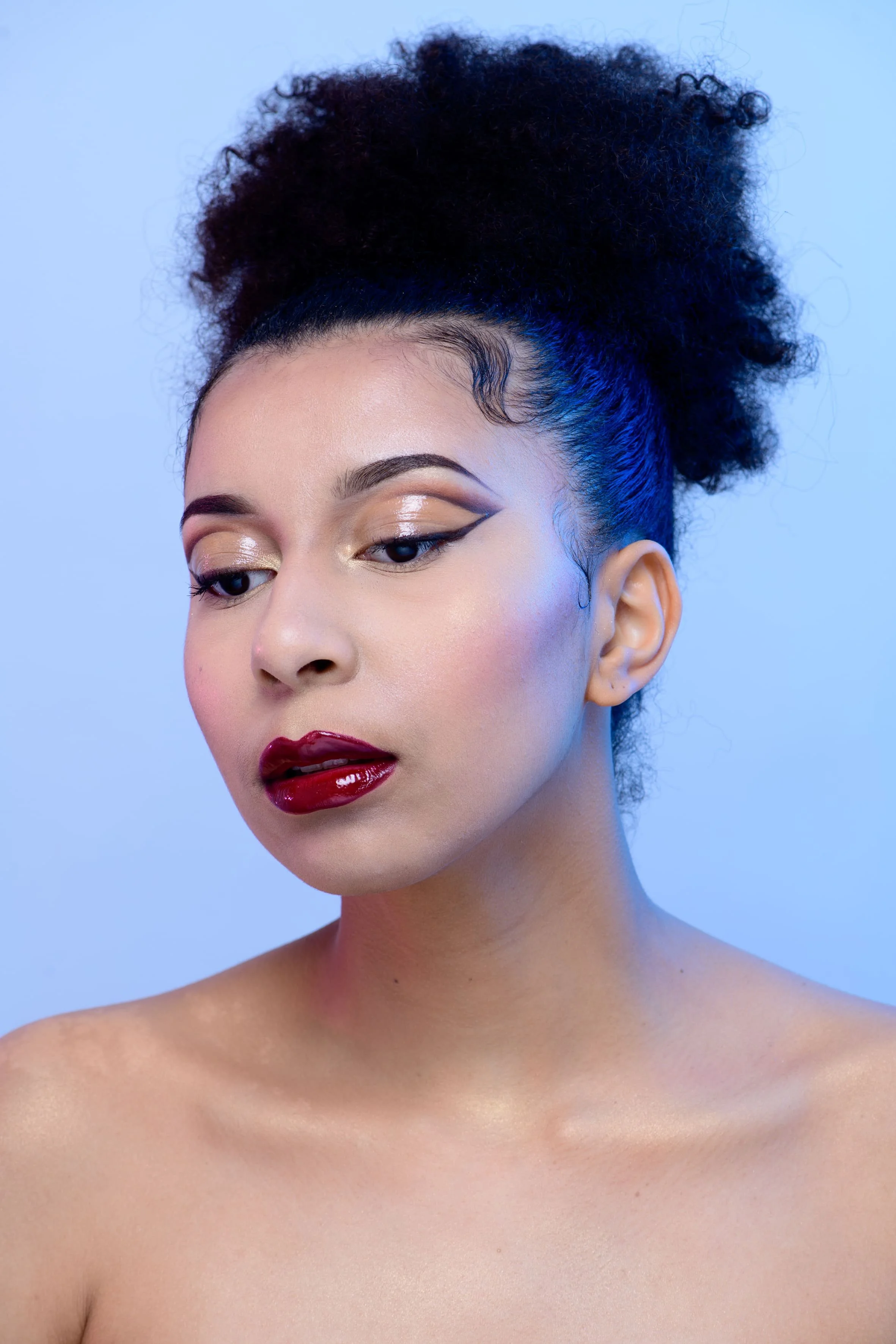 Close-up d'une femme avec maquillage sophistiqué, cheveux bouclés en chignon, fond bleu clair.