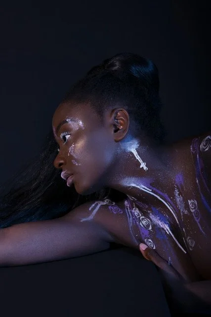 Femme avec maquillage artistique violet et blanc, posant contre un fond noir.