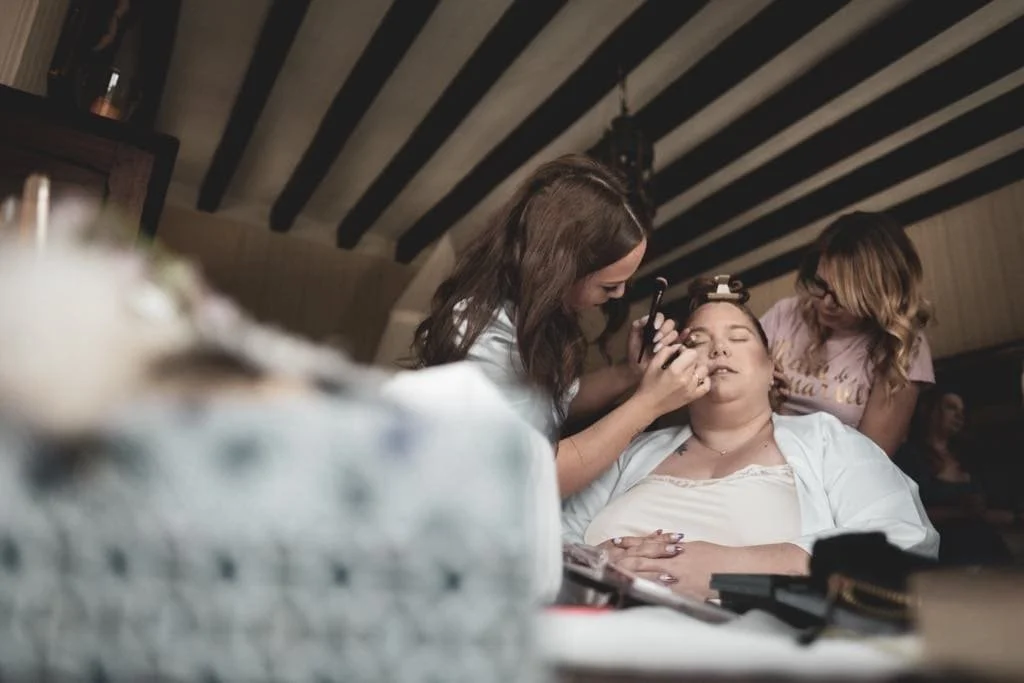 Un maquillage en train d'être appliqué à une femme allongée, avec deux autres femmes aidant, dans un intérieur avec un plafond à poutres.