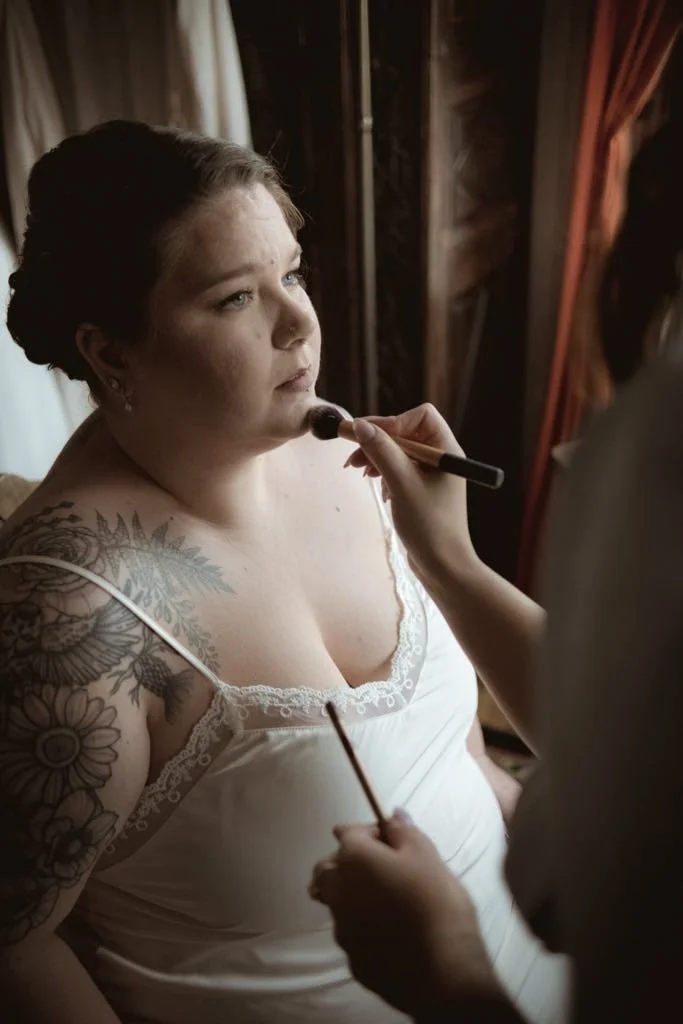Femme avec tatouages au bras gauche, en robe blanche, recevant une séance de maquillage lors d'une préparation pour un événement.