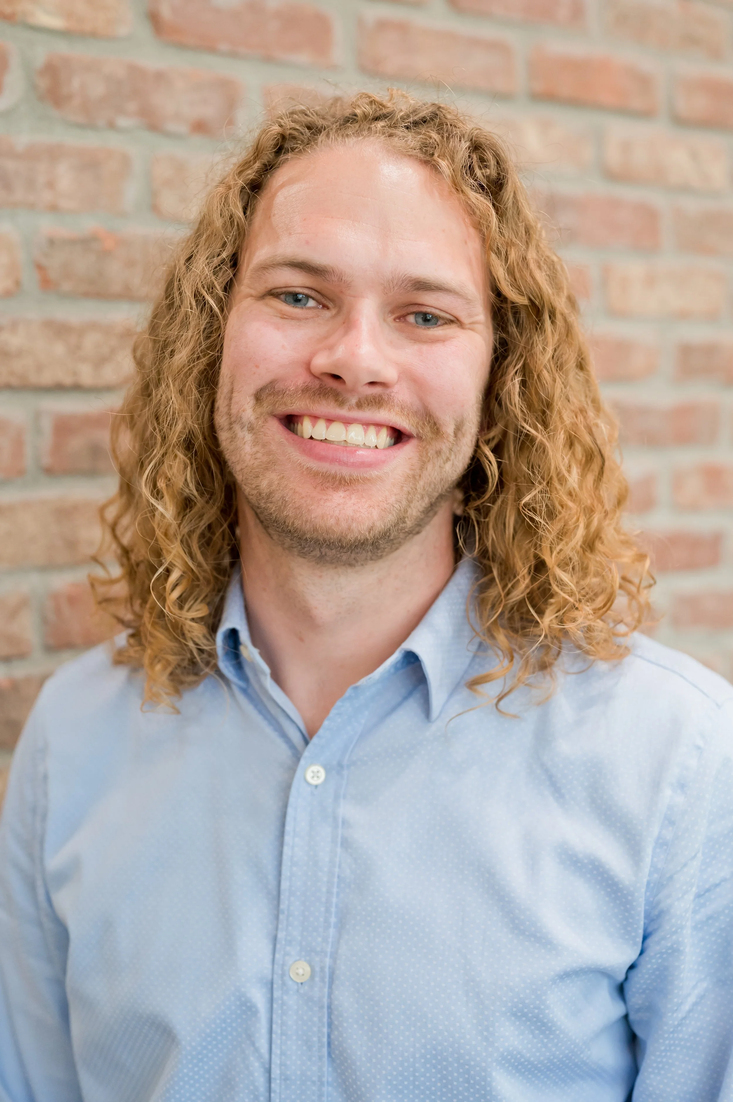 Dr. Tanner Duncan, with long, curly blond hair and a light blue button-up shirt, is smiling in front of a brick wall background.