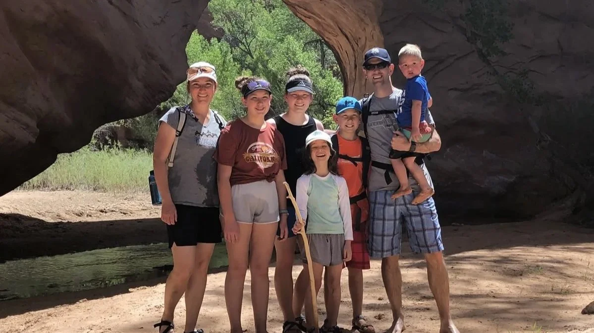Dr. Ryan Bells's family of seven, standing outdoors under a rocky natural arch. They are smiling and dressed in casual summer clothing, with some wearing hats and sunglasses, on a sunny day with greenery in the background.
